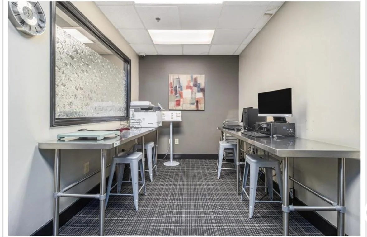 Small office or break room with two stainless steel tables, four chairs, a wall-mounted painting, and a computer station with a monitor and printer. Window shows indoor foliage outside.