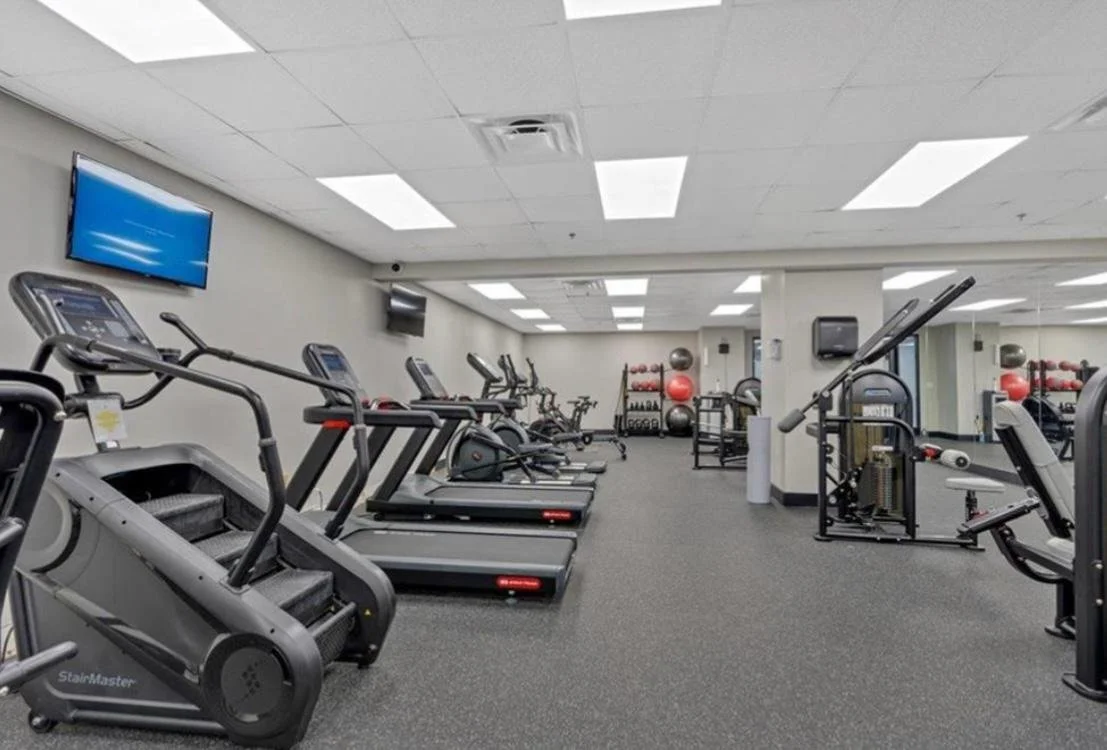A gym with elliptical and treadmill exercise machines along the left side, free weights, balance balls, and workout equipment toward the back, and a mirror wall on the right side.