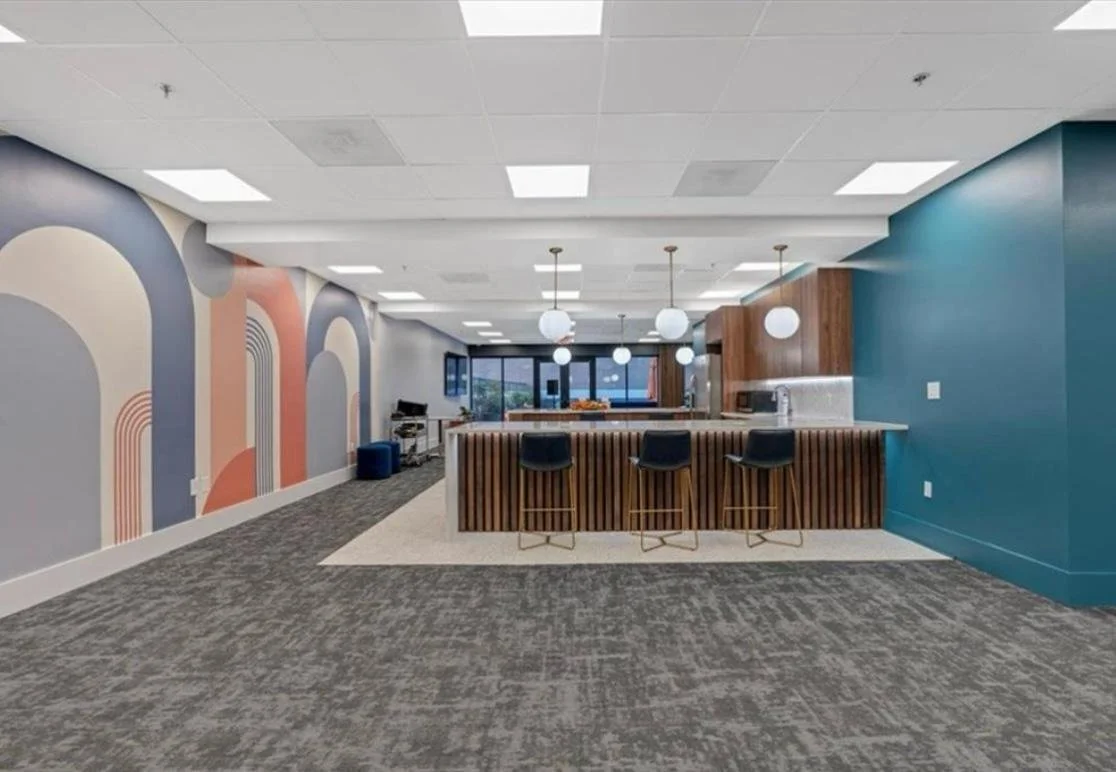 Modern office lounge with a bar area, four black chairs, colorful abstract wall mural, wooden accents, and pendant lighting.