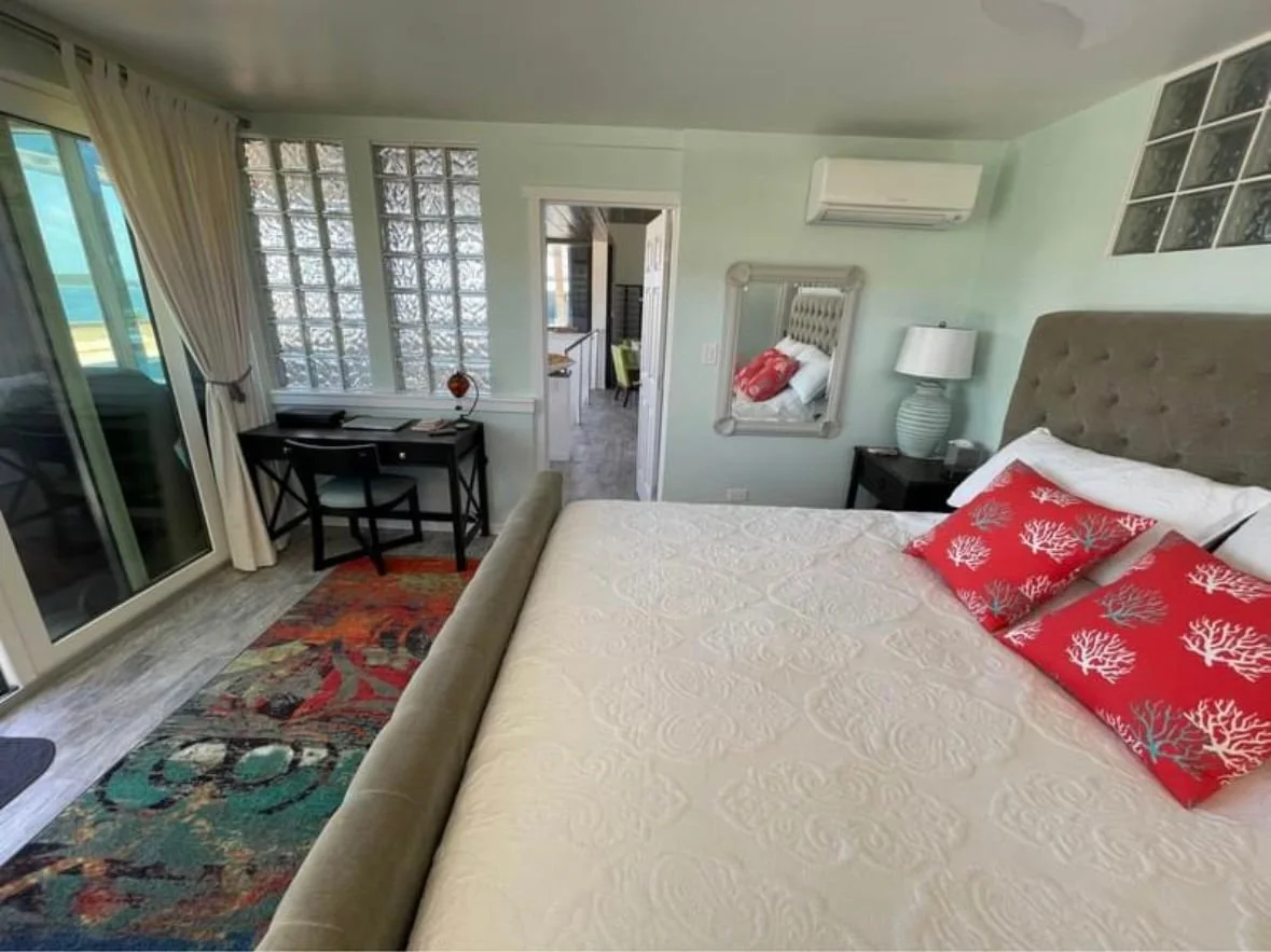 A cozy bedroom with a large bed, red coral-patterned pillows, and a tufted headboard. There is a nightstand with a lamp and mirror, a small desk with two chairs, a sliding door to a balcony, and a view of an outside ocean area.