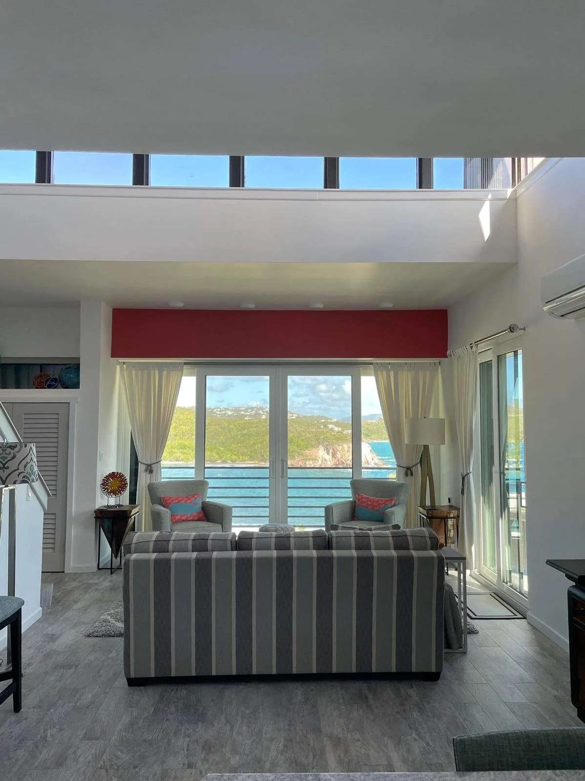 Living room with a view of the ocean through large glass sliding doors, two armchairs with pillows, a striped sofa, side tables, and a floor lamp, with a balcony railing outside