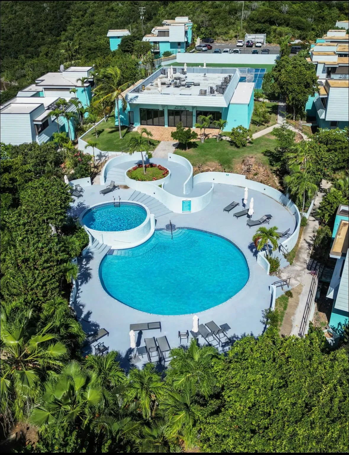 Aerial view of a modern residential complex featuring a swimming pool area with a large round pool, a smaller round hot tub, with lounge chairs and umbrellas, surrounded by lush green trees and landscaped gardens. There are multiple contemporary houses with flat roofs and outdoor seating, parked cars, and a hillside in the background.