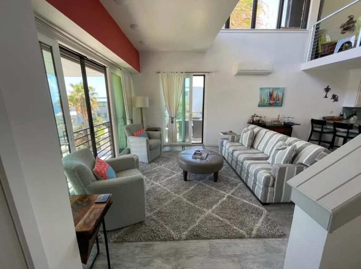 Living room with large windows, white walls, light-colored furniture, a striped sofa, two armchairs with colorful cushions, a round ottoman, and decor with tropical and sailboat themes.