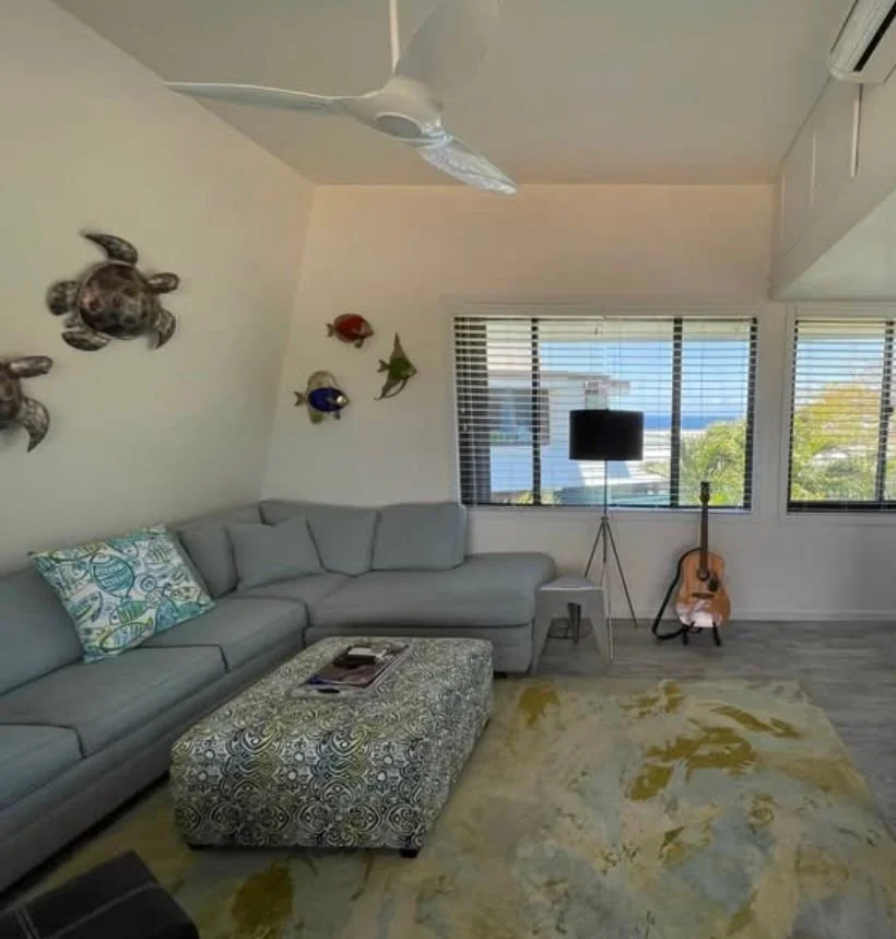 Living room with gray sectional sofa, patterned ottoman, wall-mounted turtle and fish decorations, windows with blinds, a flat-screen TV on a stand, an acoustic guitar, and a ceiling fan.