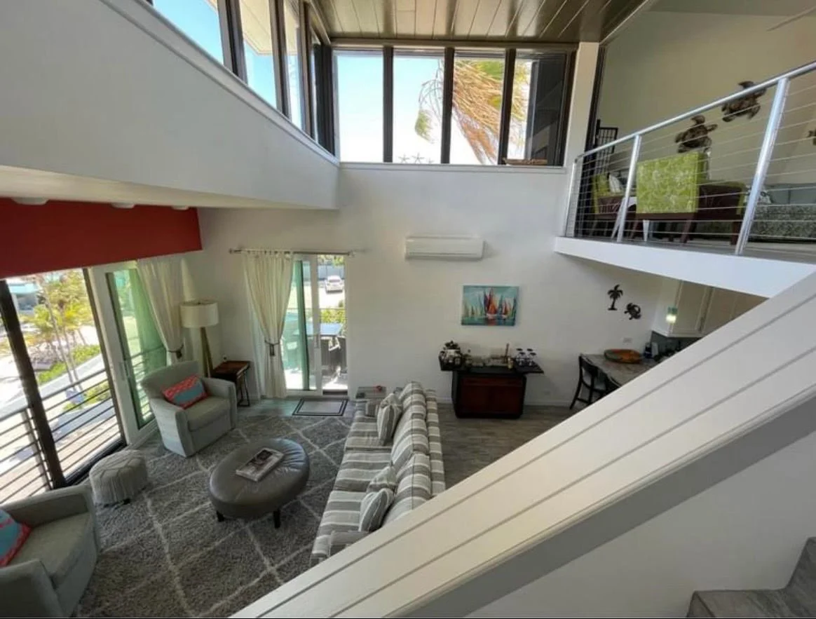 Living room with a sofa, armchairs, a round ottoman, a patterned rug, sliding glass doors opening to a balcony, large windows, and a second-floor balcony with a table and chairs.