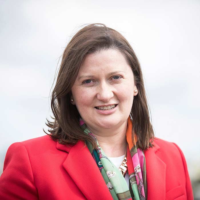 A woman with shoulder-length brown hair, wearing a red blazer and a colorful scarf, smiling outdoors.