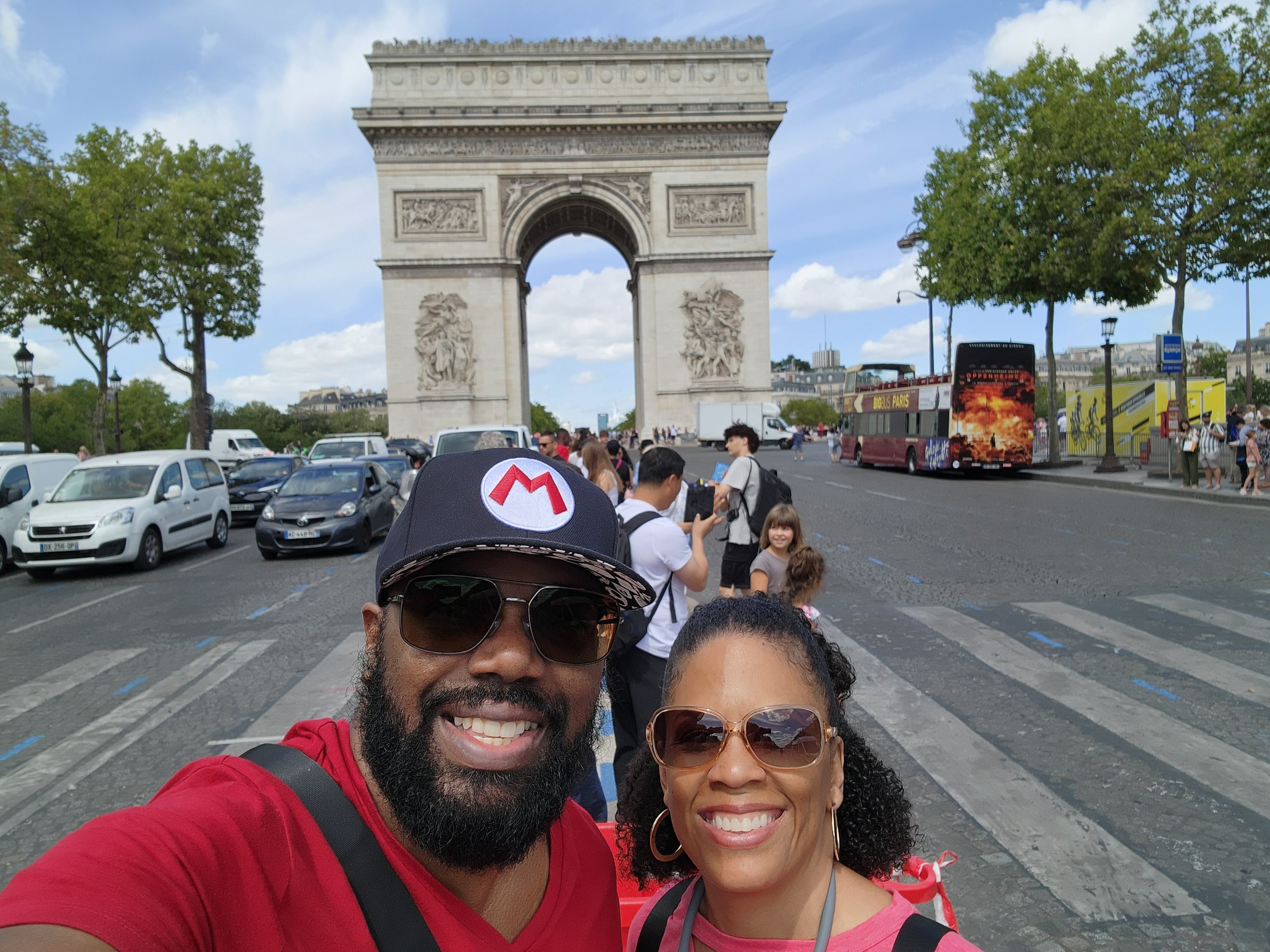 A smiling couple taking a selfie in front of the Arc de Triomphe in Paris, France on a sunny day with some tourists, cars, and a double-decker tour bus in the background.