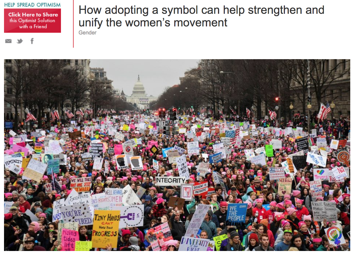 Large crowd of women participating in a march or protest, holding various signs and banners. Many women are wearing pink hats. The U.S. Capitol building is visible in the background.
