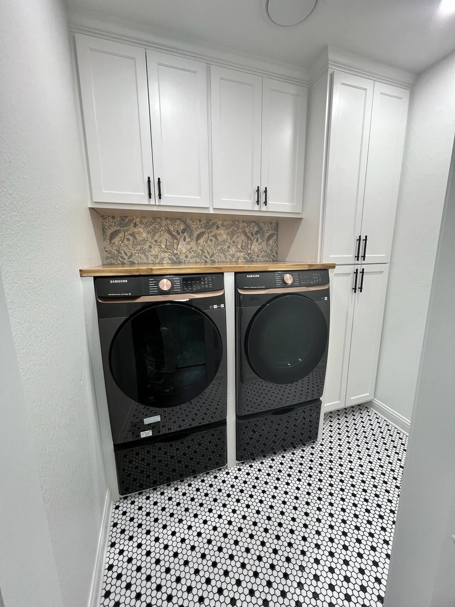 Laundry room renovation with updated black & white patterned floor tile, custom-built & painted cabinetry including tall storage for cleaning supplies, butcher board countertop & sliding door entry.