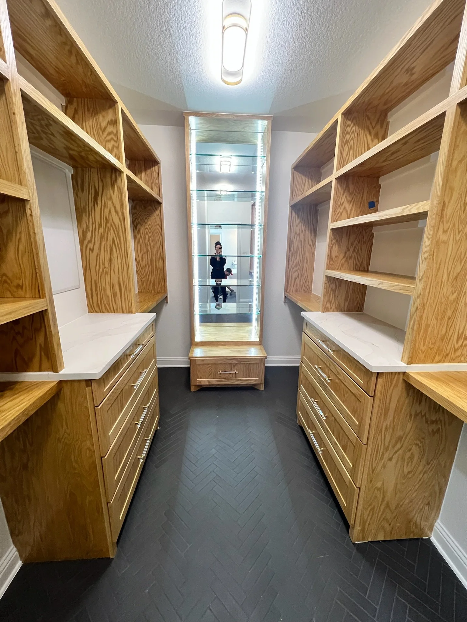 Primary closet renovation with herringbone stone tile flooring, custom built-in drawers, quartz countertops, stained red oak open shelving, glass shoe display with bench seat & storage drawer. LED lighting installed around display & in ceiling.