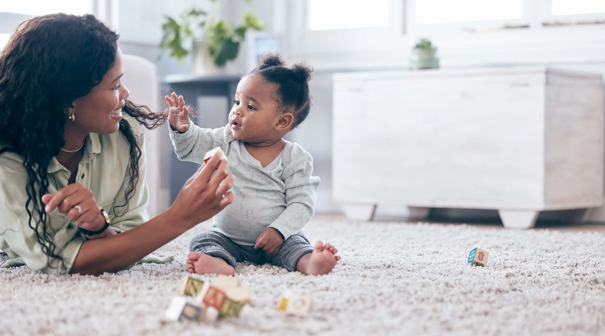 A woman and a young child playing with building blocks on a beige carpet in a bright living room.
