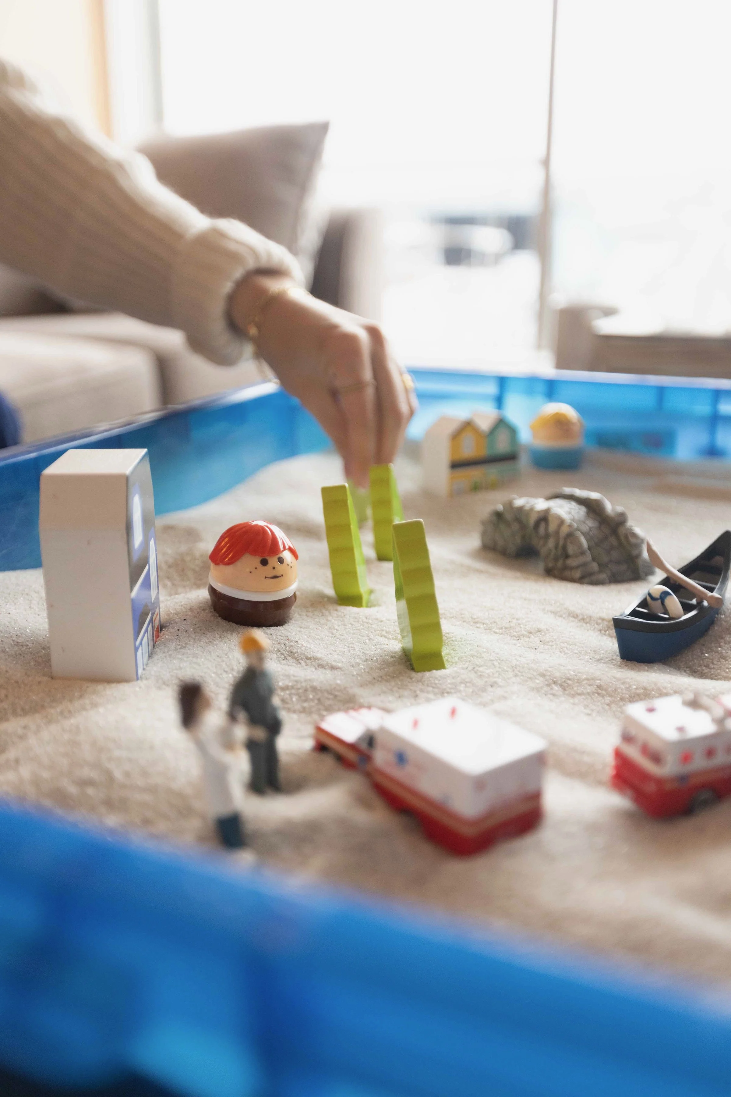 A person’s hand arranging toy figures and objects on a sandy surface, creating a miniature beach scene with a small boat, miniature buildings, and figurines.
