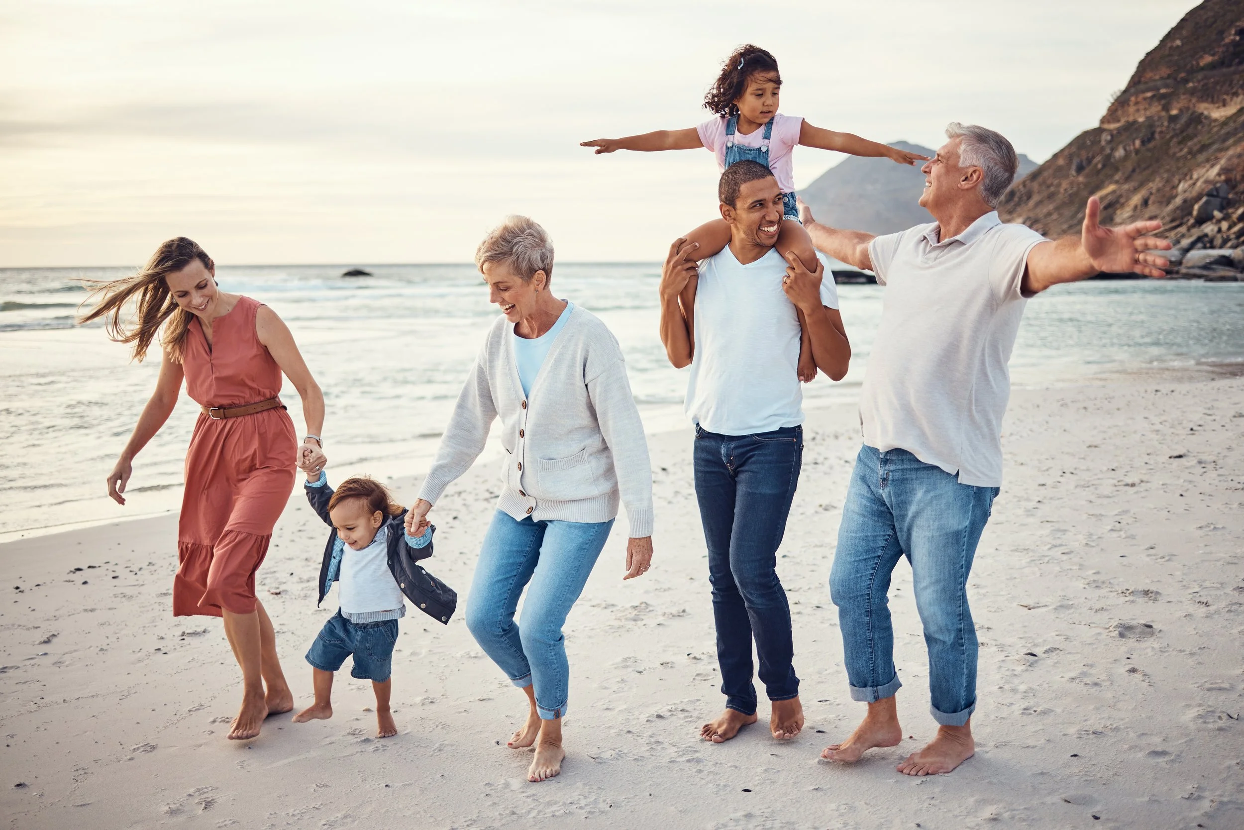Family of six enjoying a walk on the beach, with a young girl riding on a man's shoulders, during sunset.