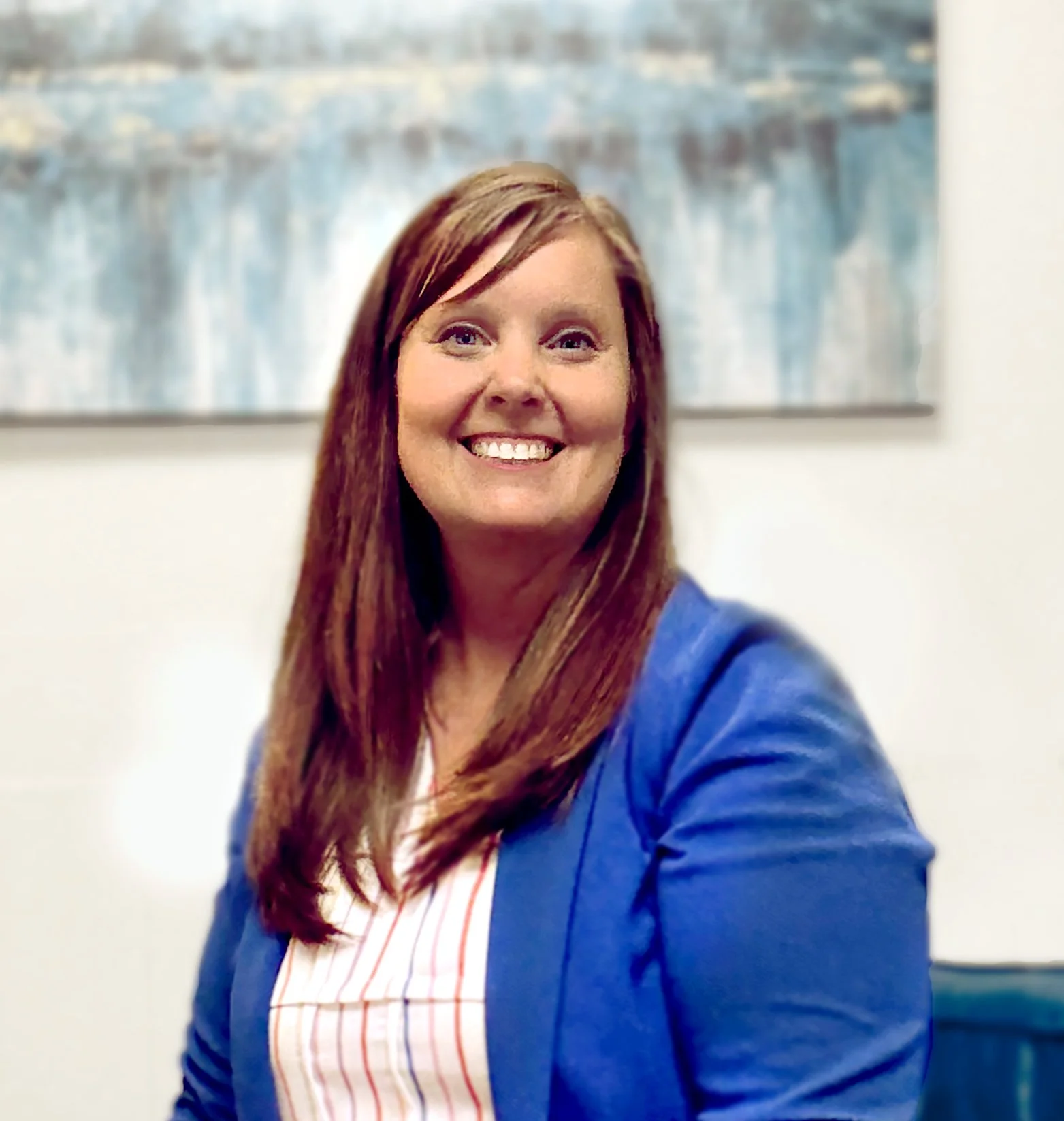 Woman with long red hair smiling, wearing a blue blazer and a white blouse with red stripes, sitting in front of abstract art on a white wall.