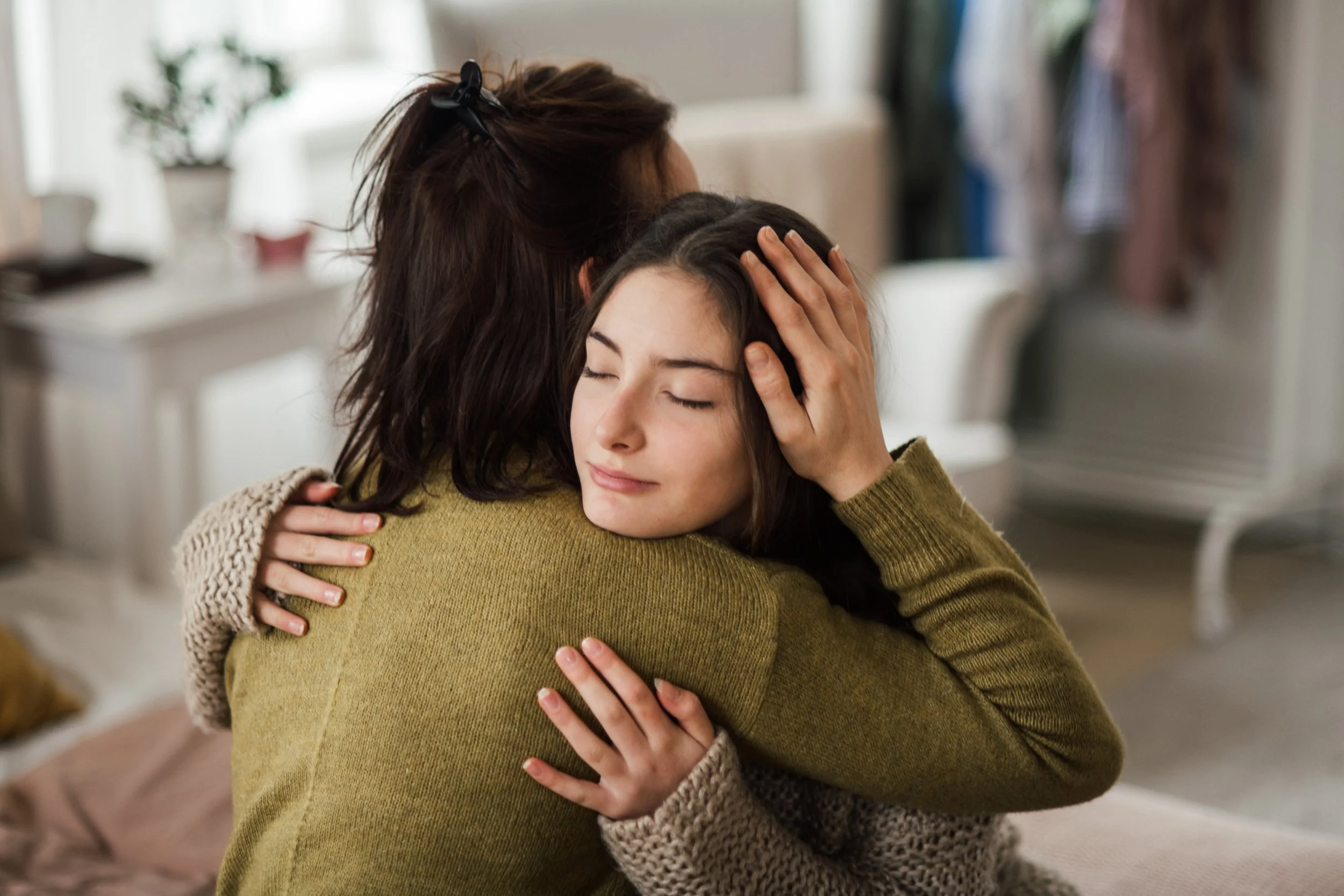 Two women hugging each other warmly, with one leaning into the other's embrace, eyes closed, in a cozy, well-lit room.