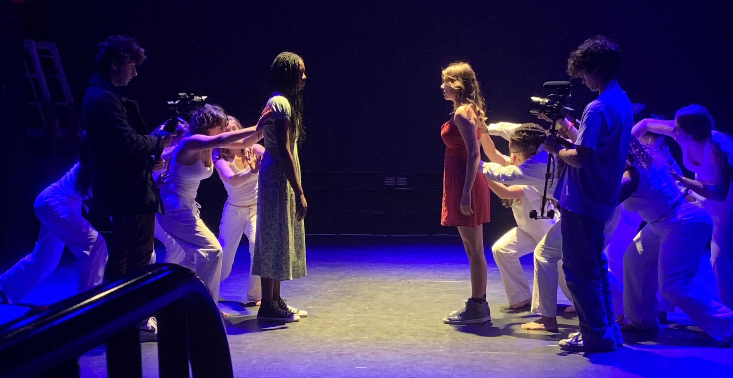 The scene shows a theatrical performance or rehearsal with two women in the center, facing each other. Several crew members and performers surround them, some holding cameras and equipment. The stage is dimly lit with blue and purple lighting, highlighting the two women in the middle.