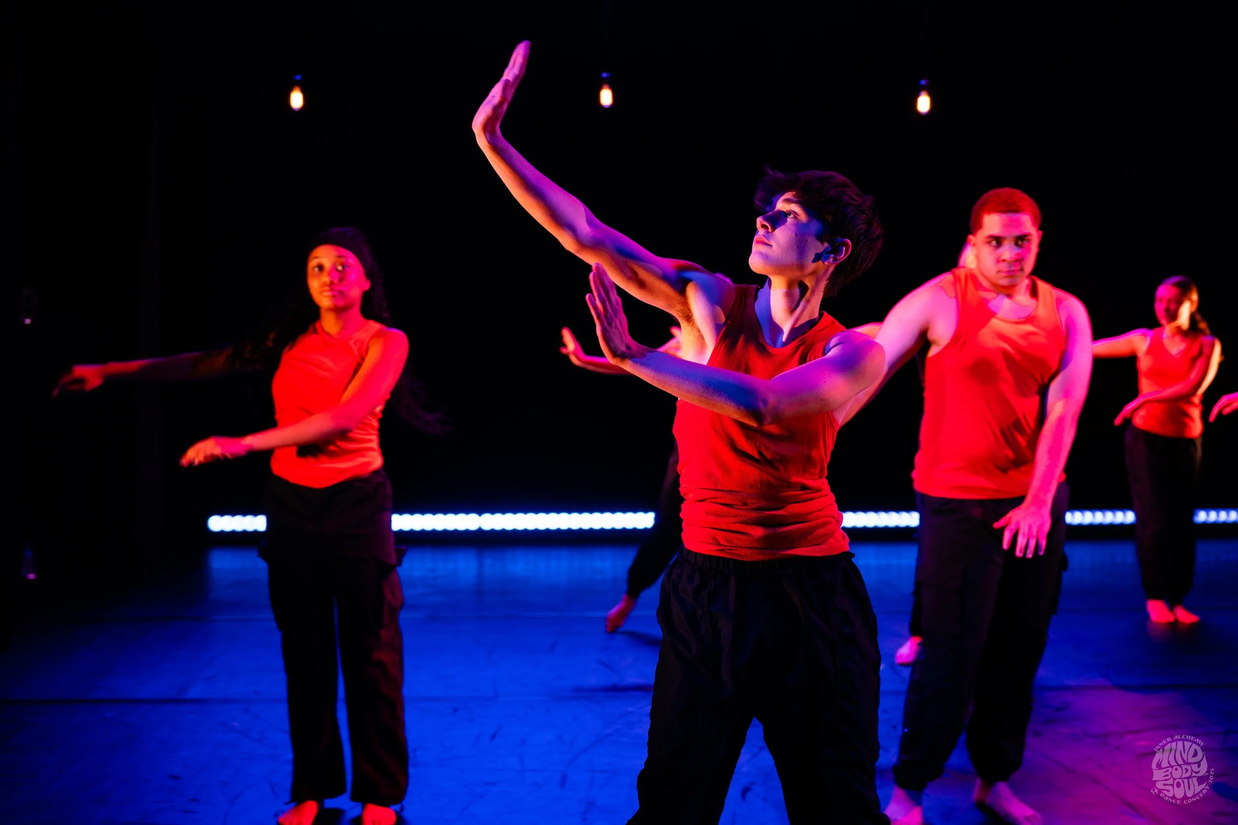 Group of diverse dancers performing on stage in red tops and black pants, with colorful stage lighting and dark background.