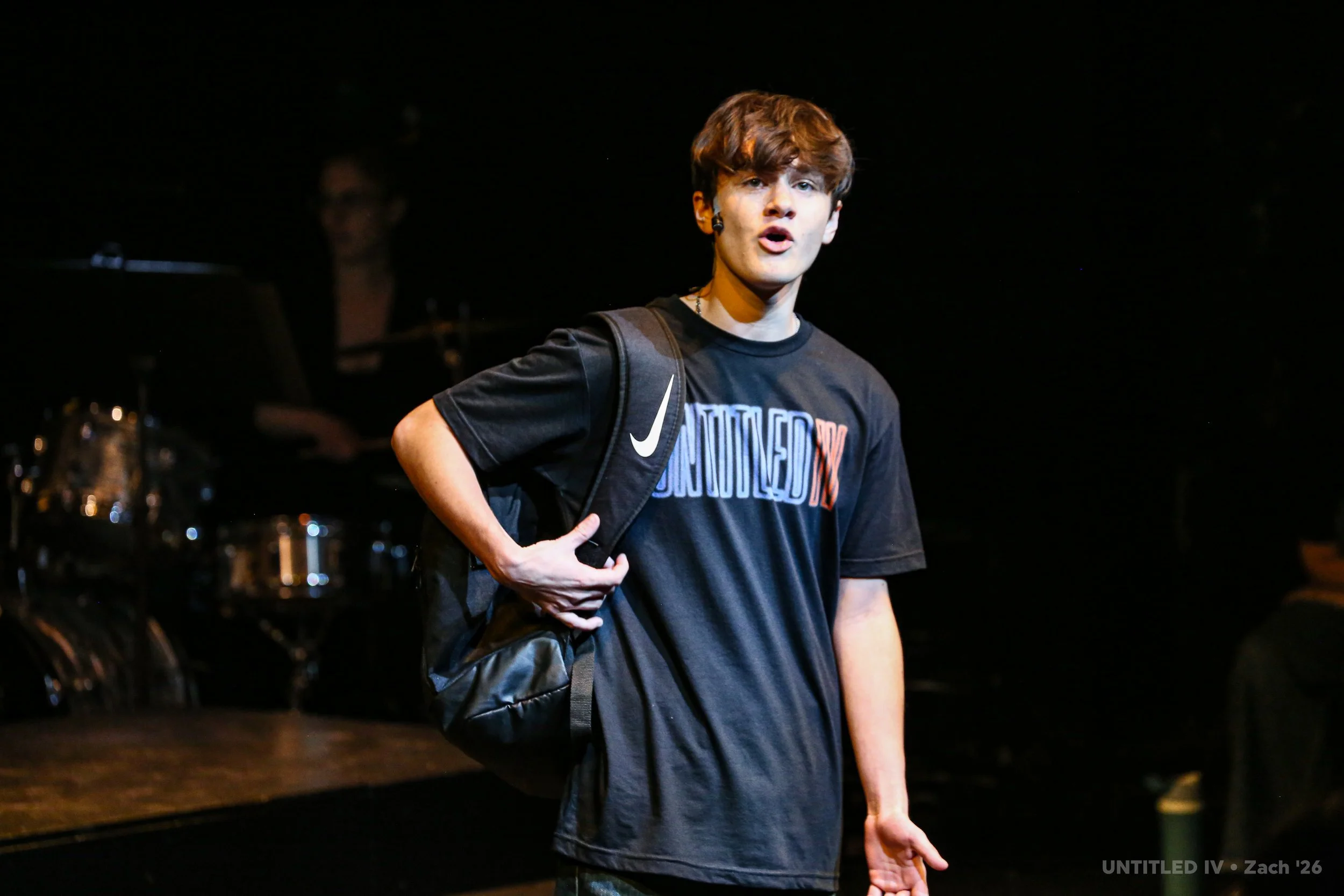 A young man with brown hair wearing a black T-shirt with a colorful print, carrying a black Nike backpack, stands on a dark stage with a drum set in the background.
