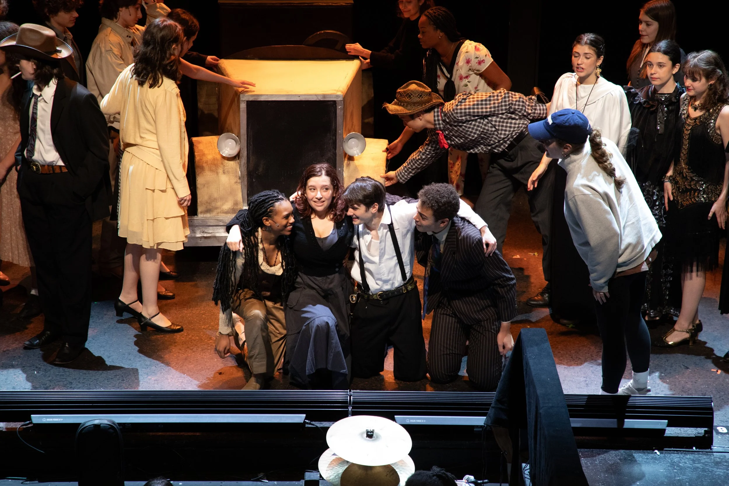 Actors performing on stage in a theatrical production, with some actors kneeling and embracing in the center and others standing around them, all in costume.