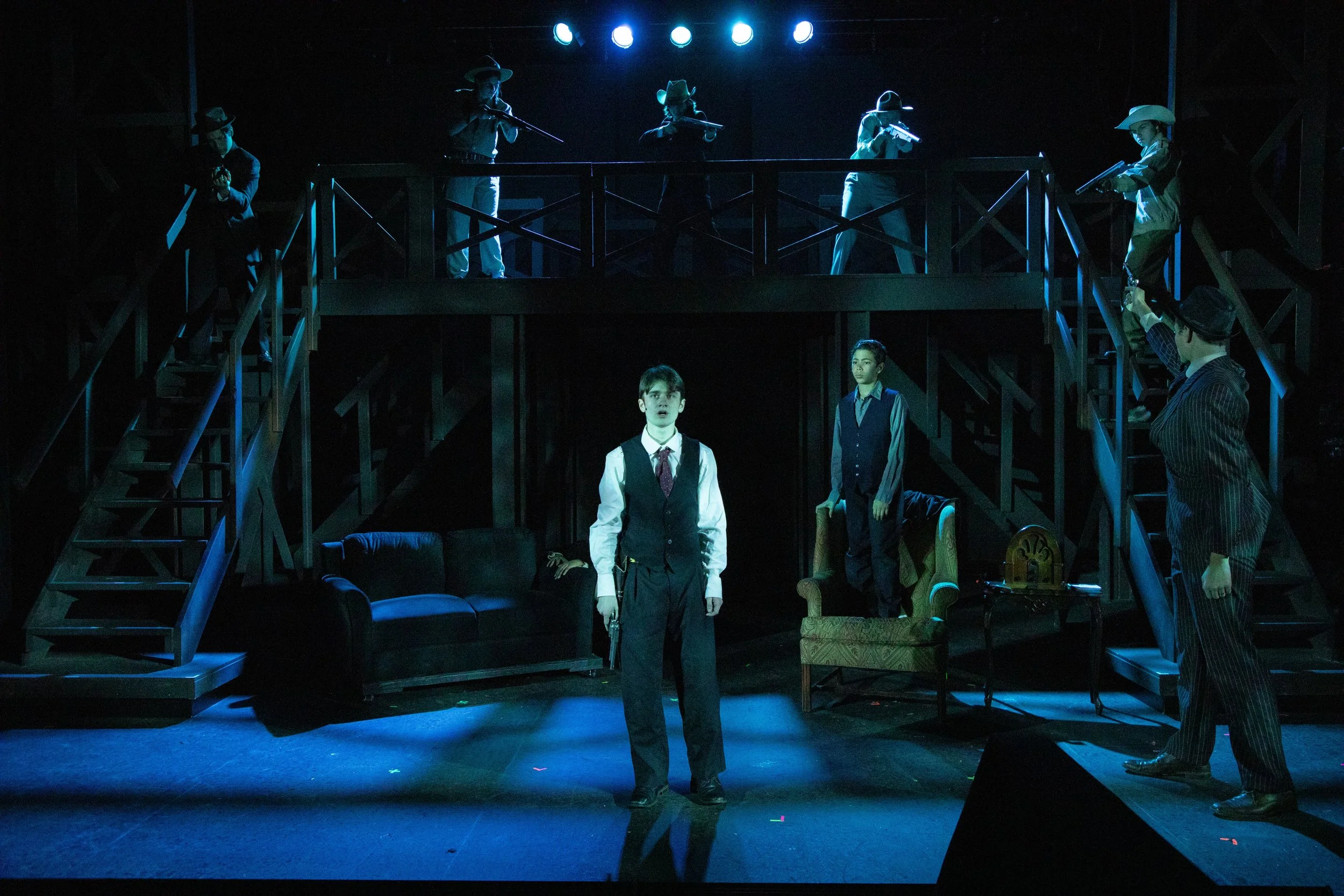 A theatrical stage scene with actors and figurines, including a young man in a vest and tie in the center, and other characters and figurines dressed in period costumes on a two-tiered set. Stage lights cast blue hues over the scene.