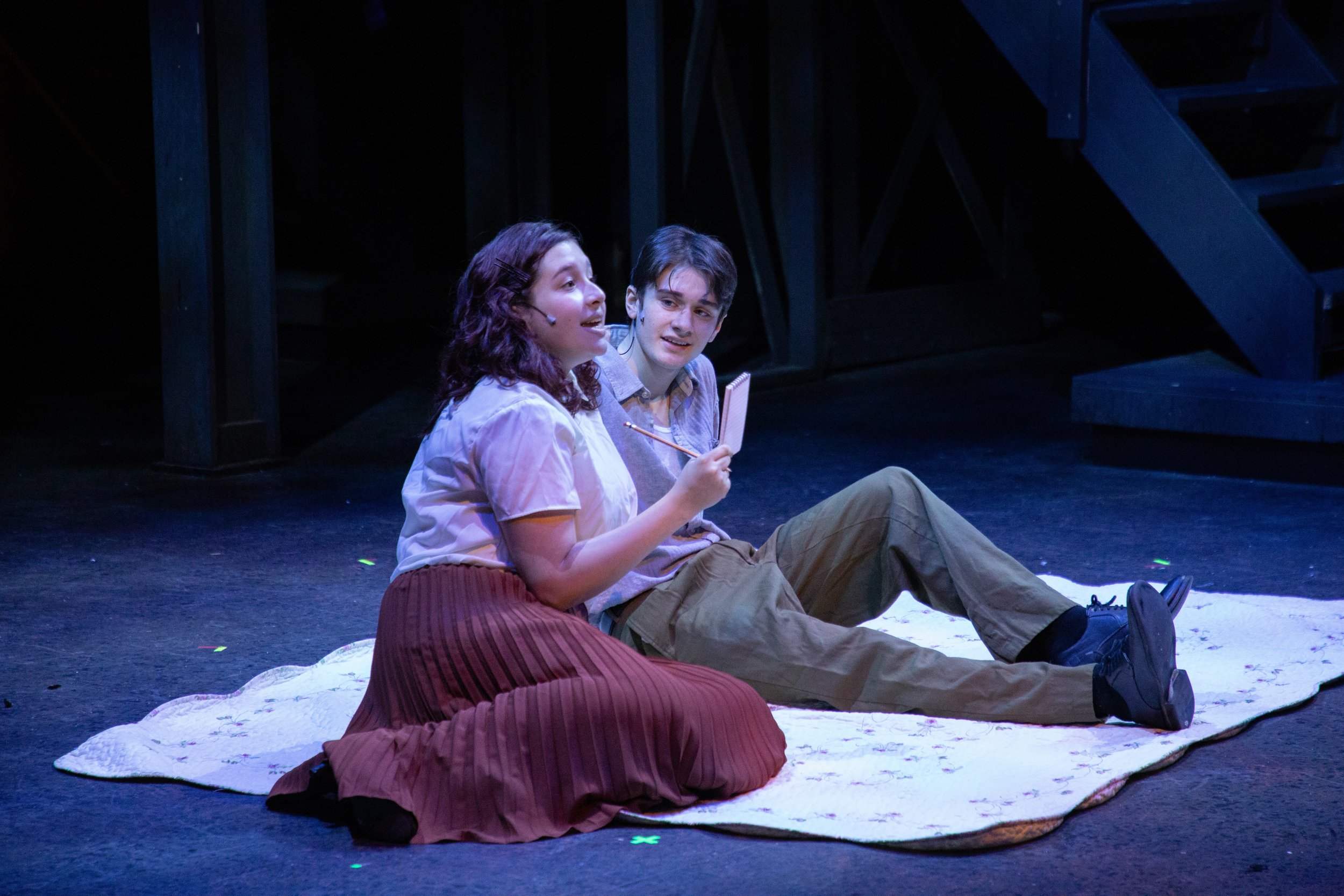 Two actors sitting on a white blanket on stage, performing in a theater, with dark background and stage stairs in the background.