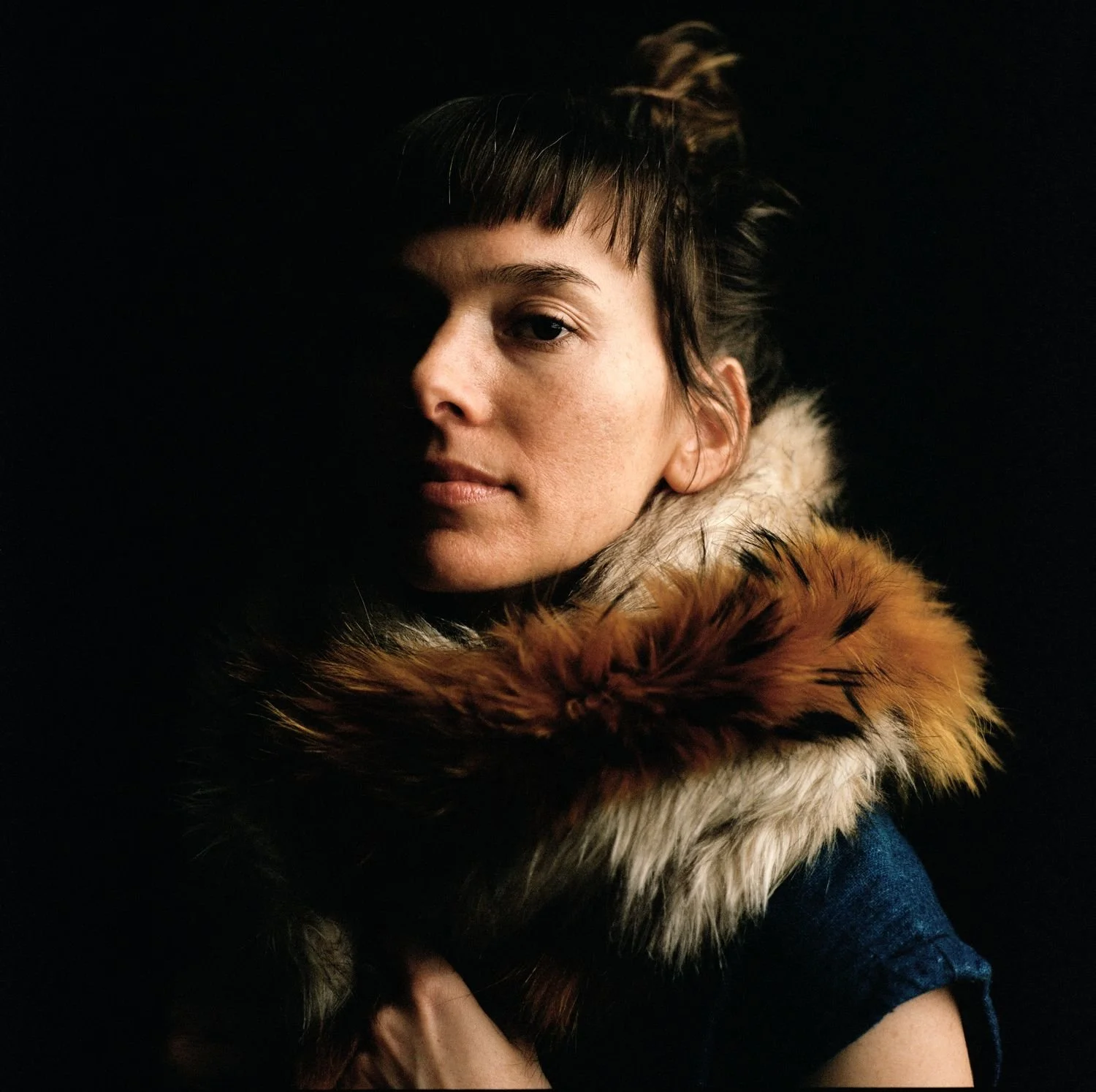 Portrait of a woman with short brown hair and bangs, wearing a coat with a fur collar, against a dark background.