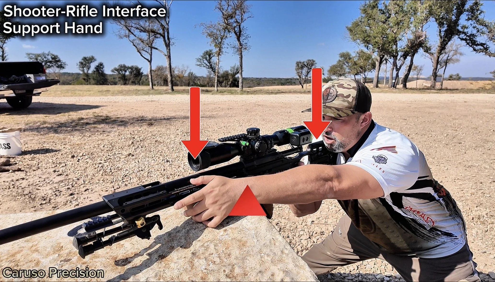 A man wearing a camouflage cap and a black and white shirt is aiming a sniper rifle with a scope supported by a rifle interface support hand on a rocky surface in an outdoor shooting range with trees and a clear blue sky in the background.