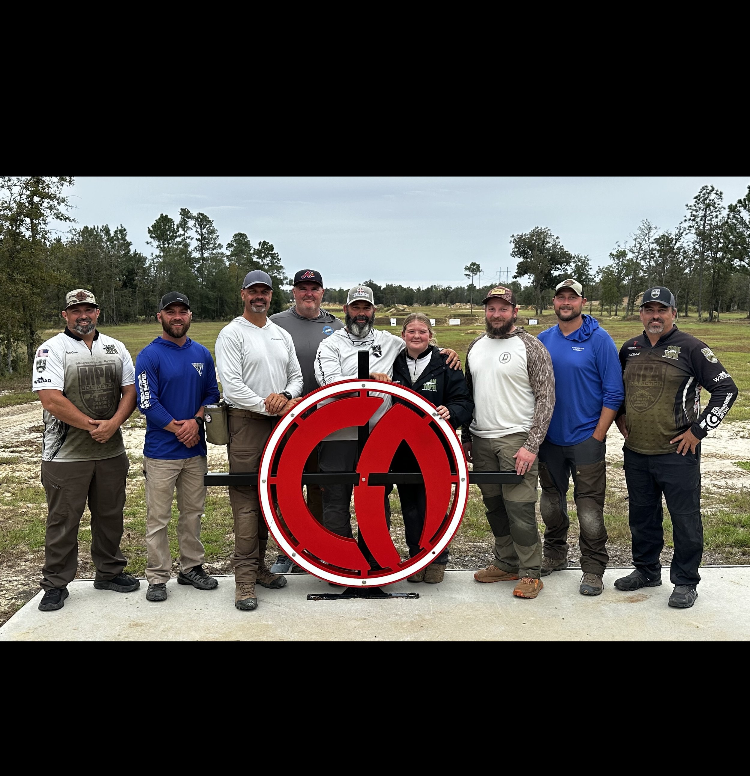 Group of nine people standing outdoors on a cloudy day, posing behind a large red and black target sign at a shooting range, with trees and open land in the background.