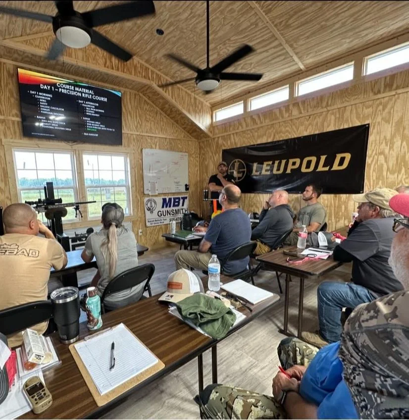 People attending a firearms training class in a wooden room with a Leupold banner, a whiteboard, and a mounted rifle on a table.
