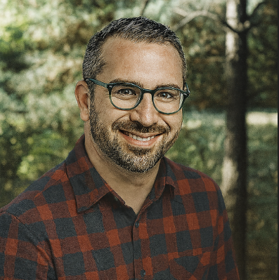 A man with short dark hair, glasses, and a beard, smiling outdoors in front of trees, wearing a red and black checkered shirt.