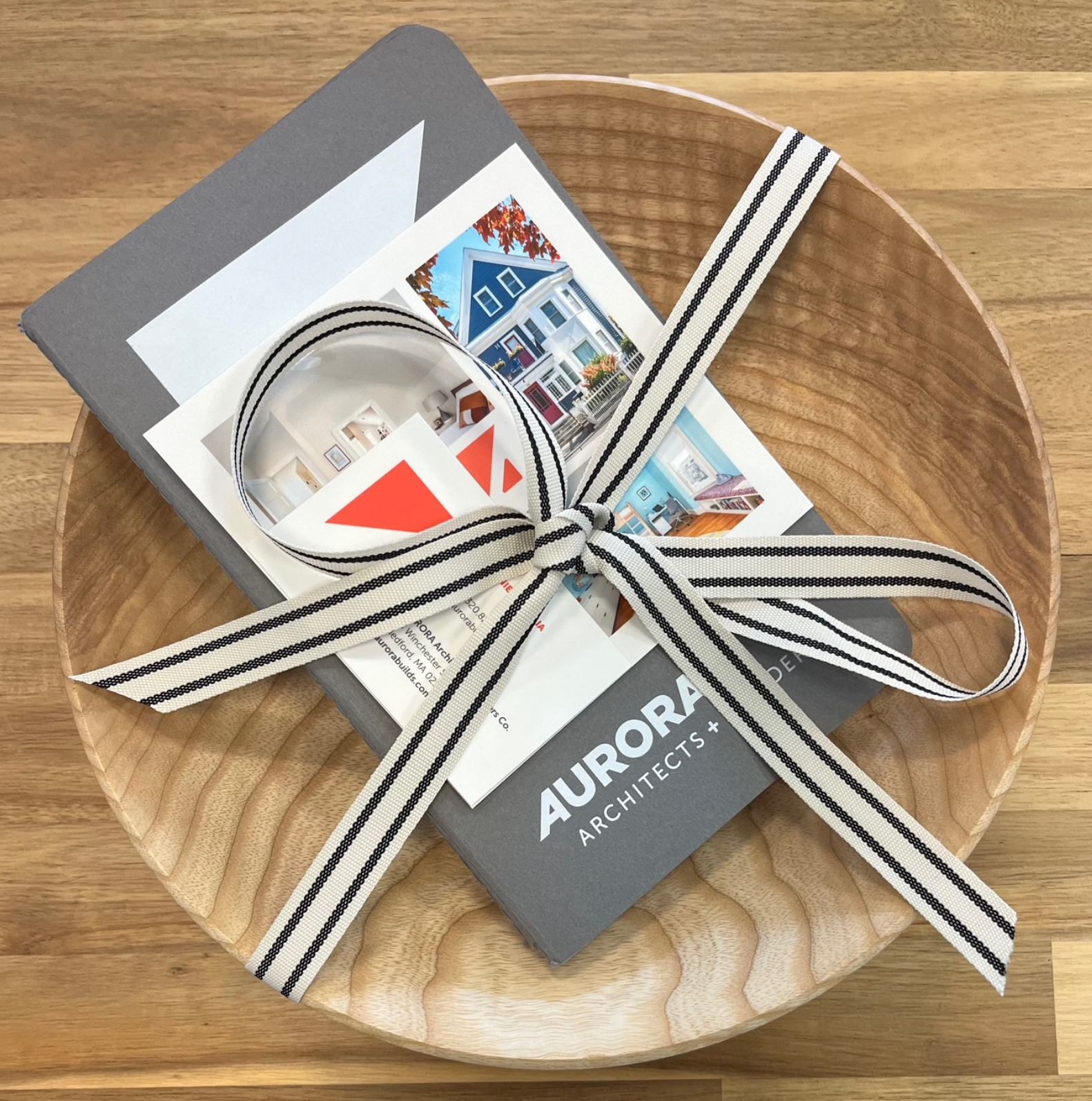 A wooden tray contains a gray notebook, vacation photos, a brochure for Aurora Architects, and a rolled striped ribbon tied in a bow.