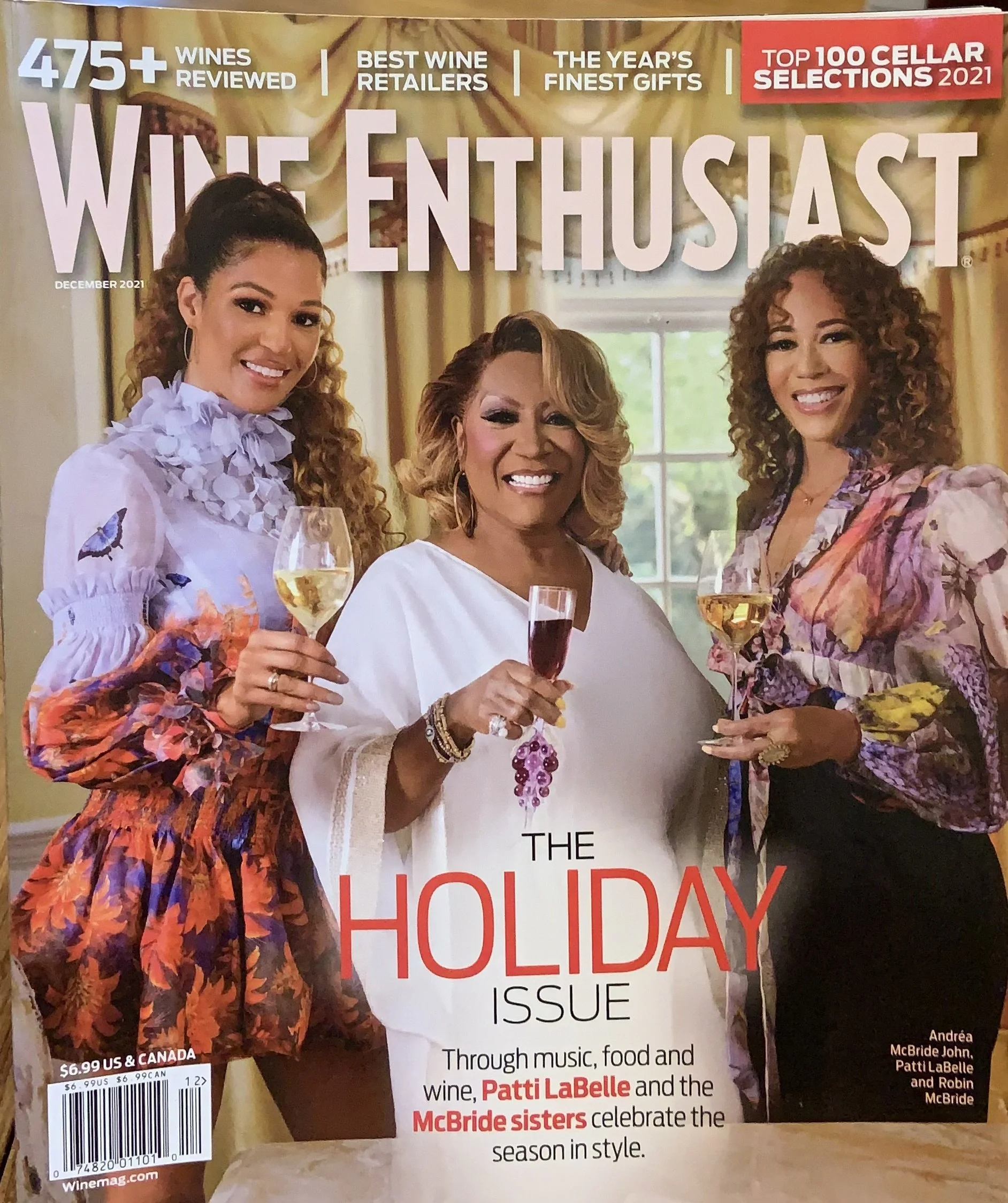 A magazine cover featuring three women smiling and holding glasses of wine inside a room with large windows. The woman on the left wears a floral dress with butterfly patterns, the woman in the middle wears a white top, and the woman on the right wears a colorful floral top. The magazine highlights include reviews of over 475 wines, best wine retailers, top gift picks, and a holiday issue celebrating music, food, and wine with the McBride sisters and Patti LaBelle.