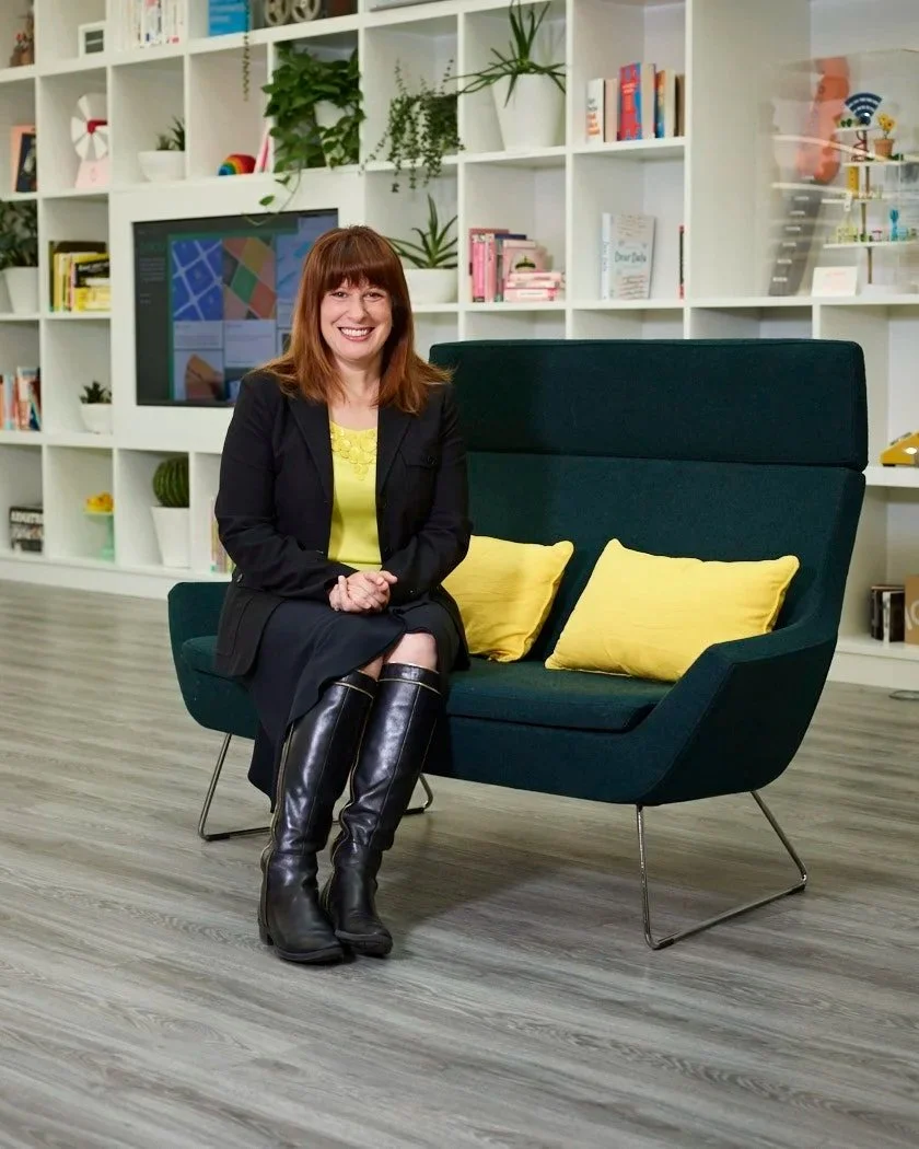 A woman with red hair sitting on a dark green modern sofa in a bright room with white bookshelves, green plants, and a flat-screen TV in the background.