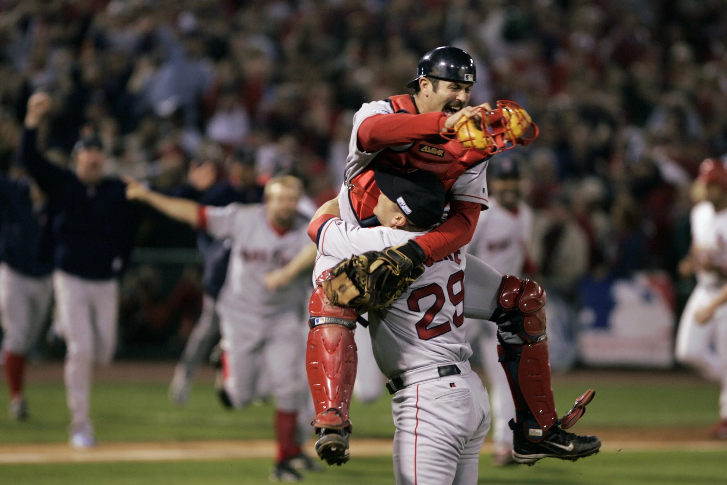 Two baseball players in team uniforms celebrating on the field, one lifting the other in a joyful embrace, with a crowd of fans in the background.