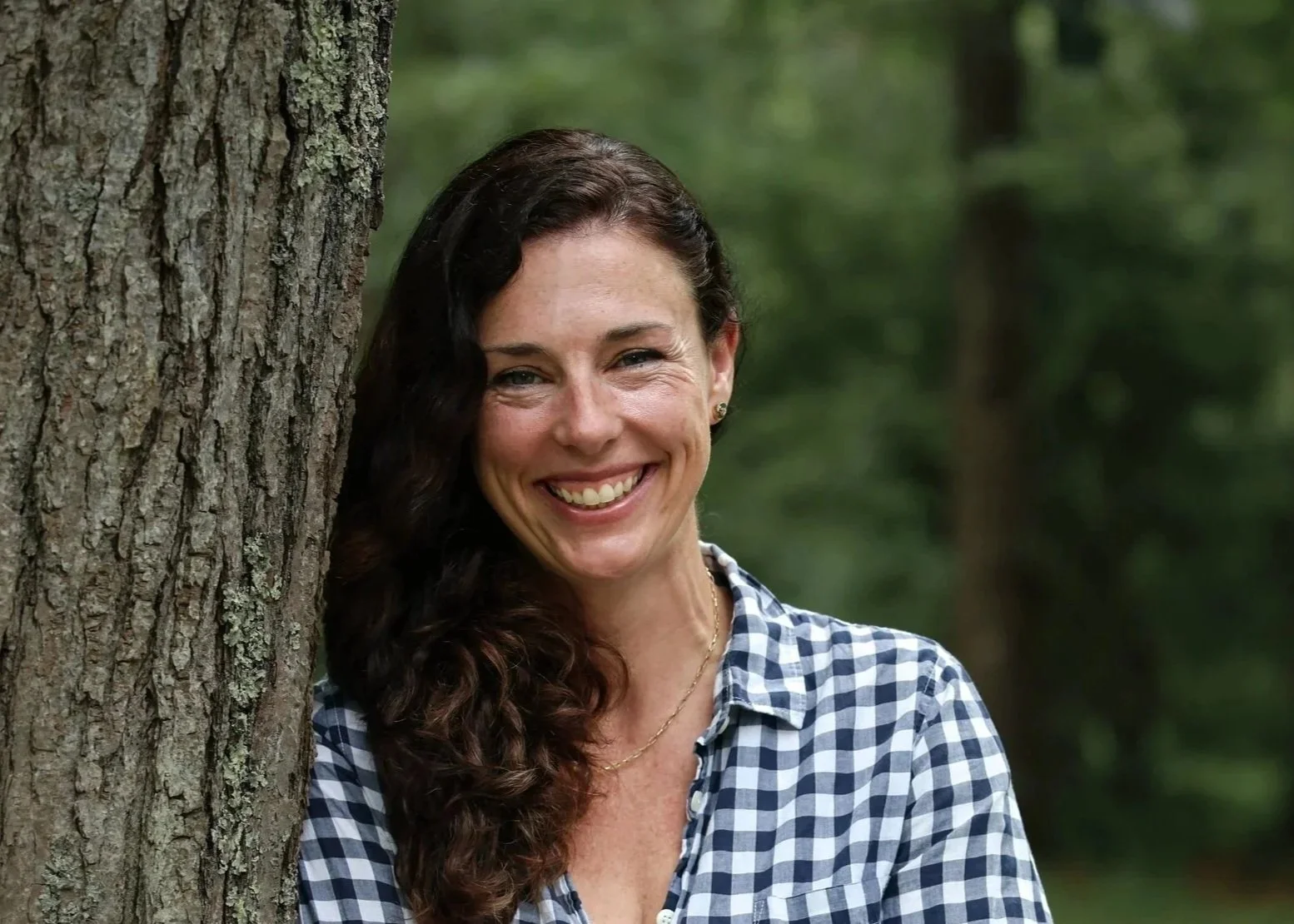 A smiling woman with long dark curly hair leaning against a tree in a forest.