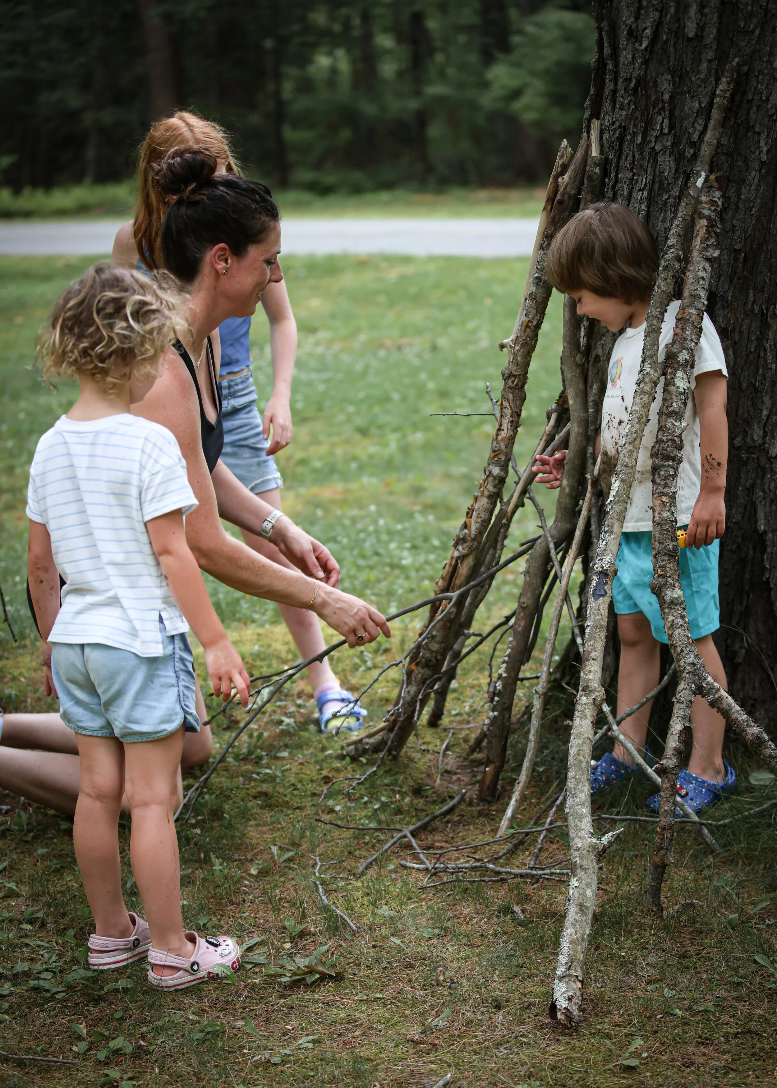 A woman and three children building a tent or fort using tree branches and sticks outdoors in a wooded area.