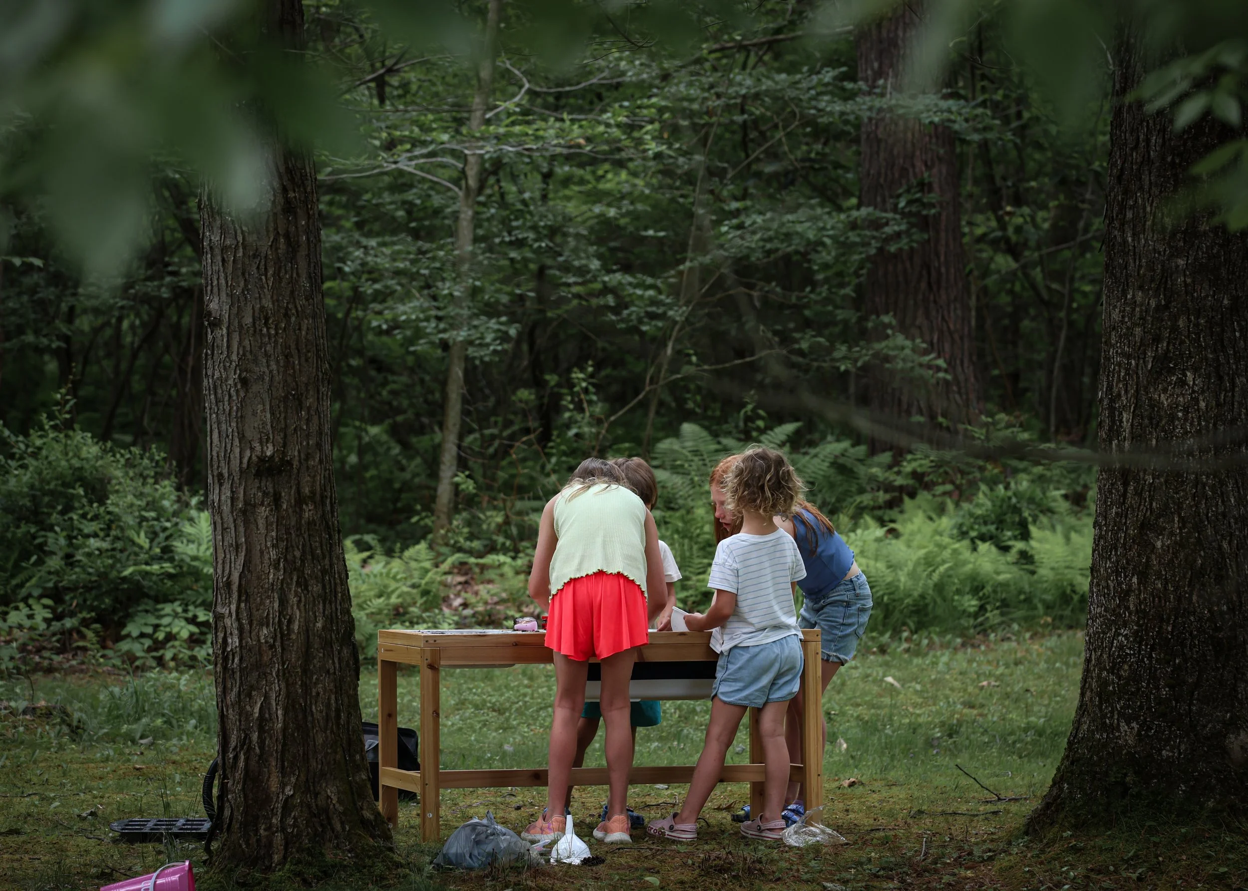 Four children standing around a wooden table in a forest, engaged in an activity, surrounded by trees and greenery.