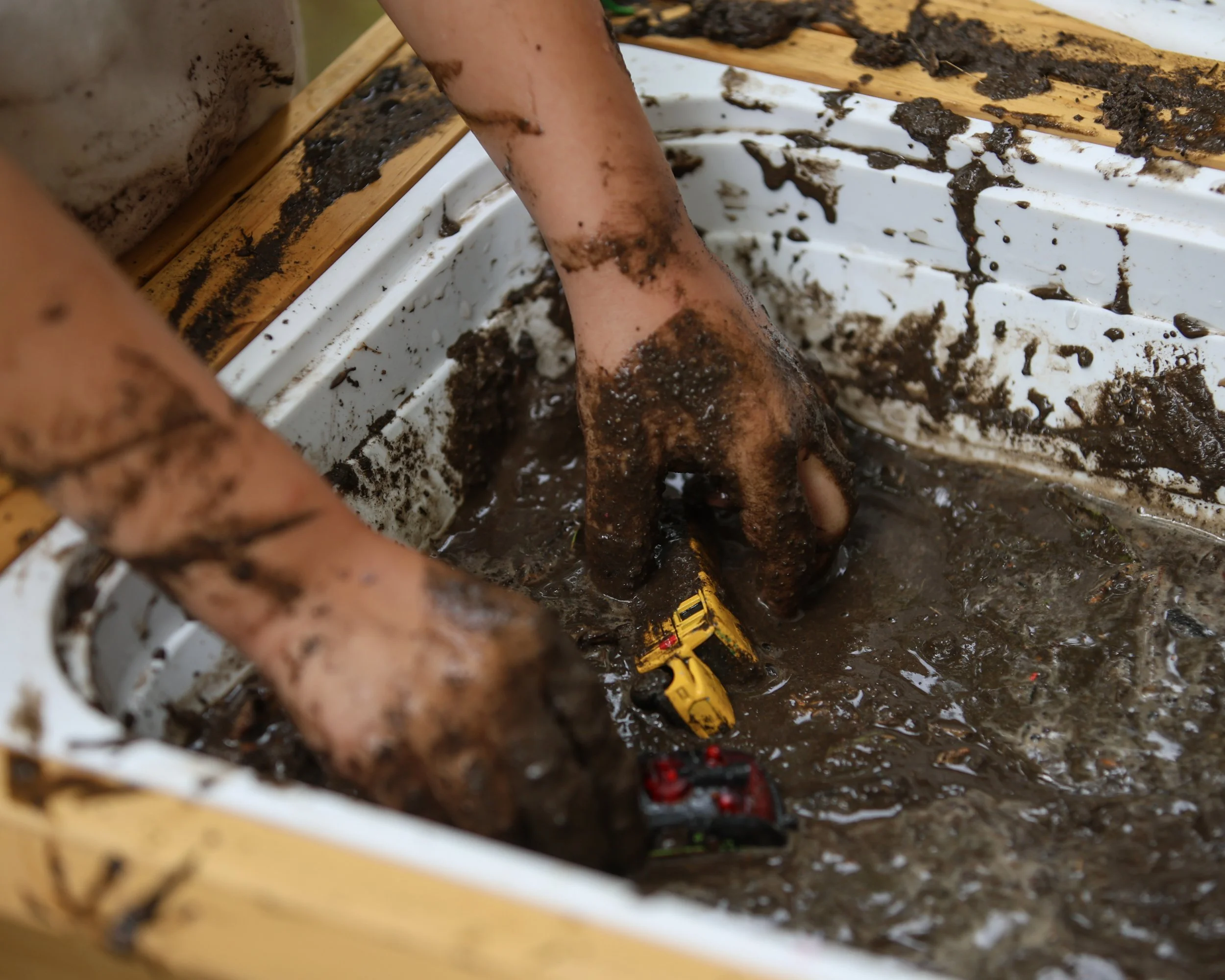 Hands playing with muddy toy construction vehicles in a muddy container.