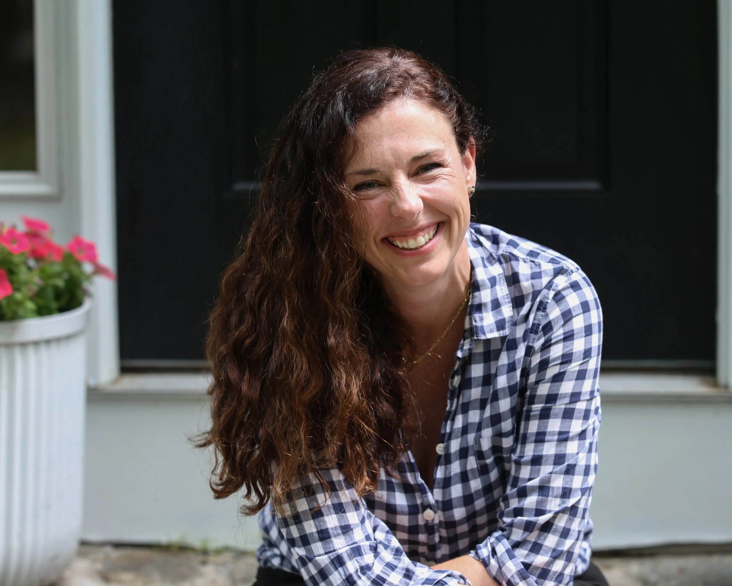 A woman with long curly brown hair wearing a blue and white checkered shirt, smiling outdoors in front of a black door and pink flowers in a white pot.