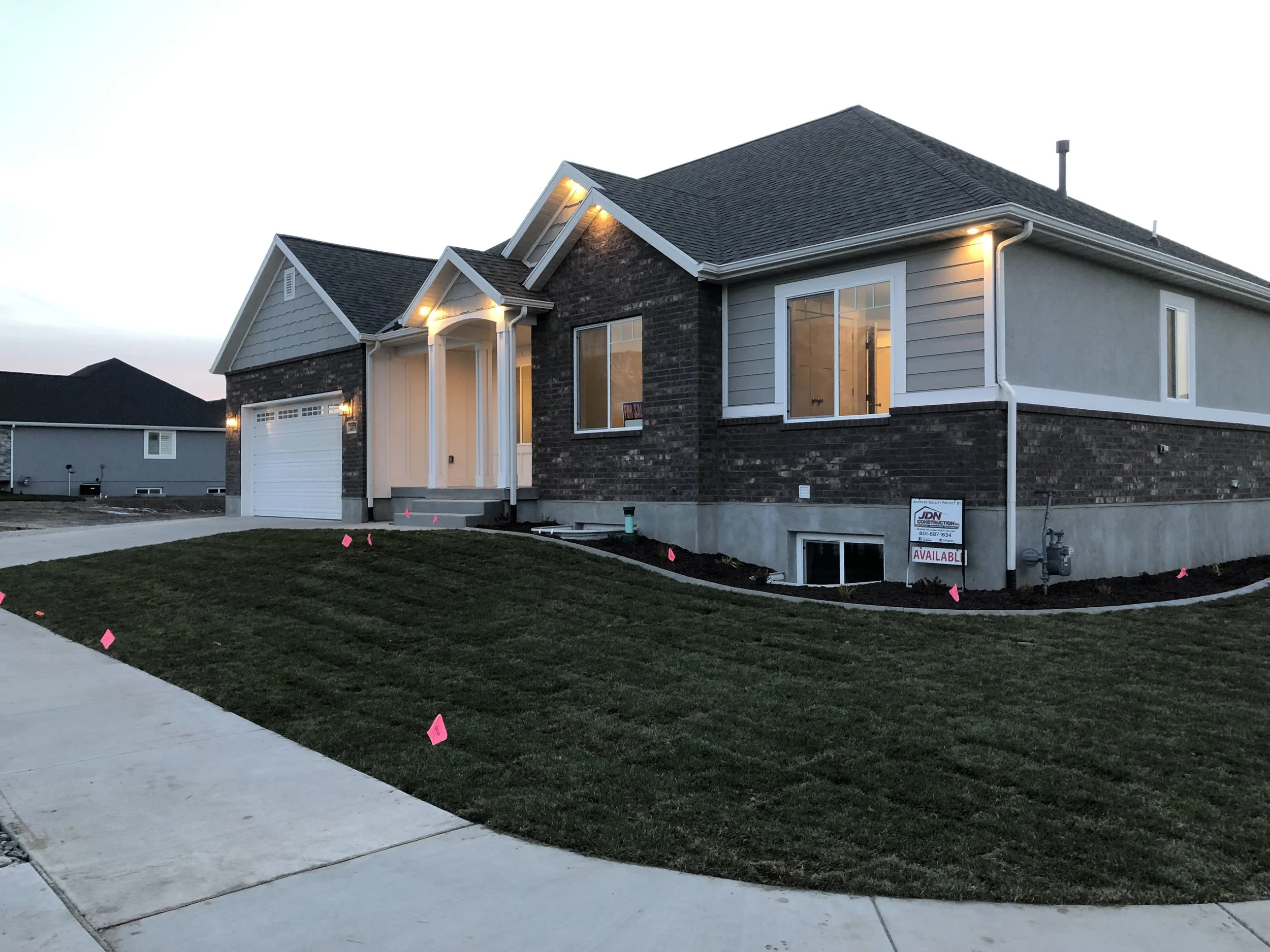 Newly constructed house with front lawn, garage, and exterior lighting visible at dusk.