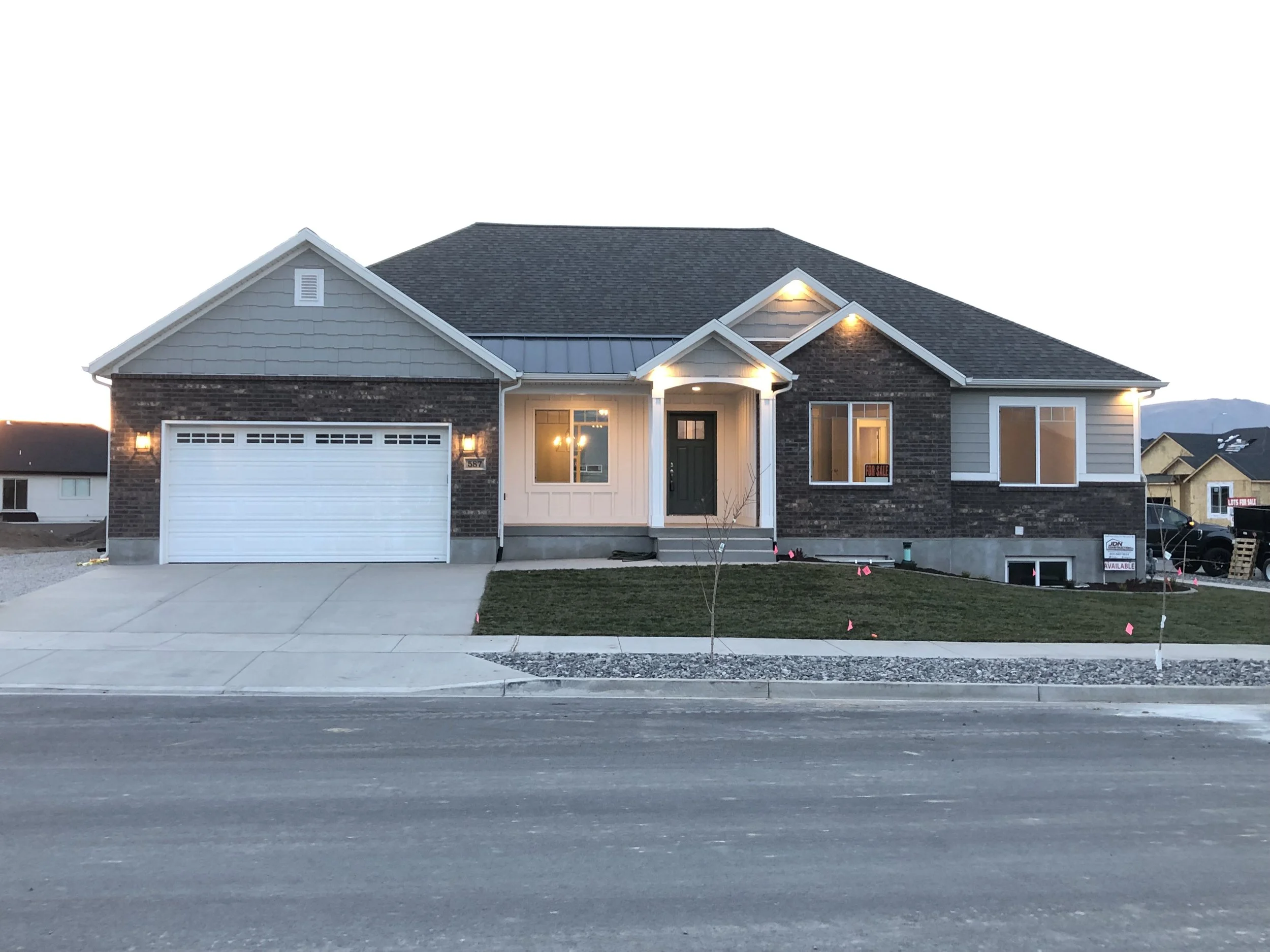 Newly built house with a two-car garage, front yard with small tree and ongoing landscaping, and exterior lights on