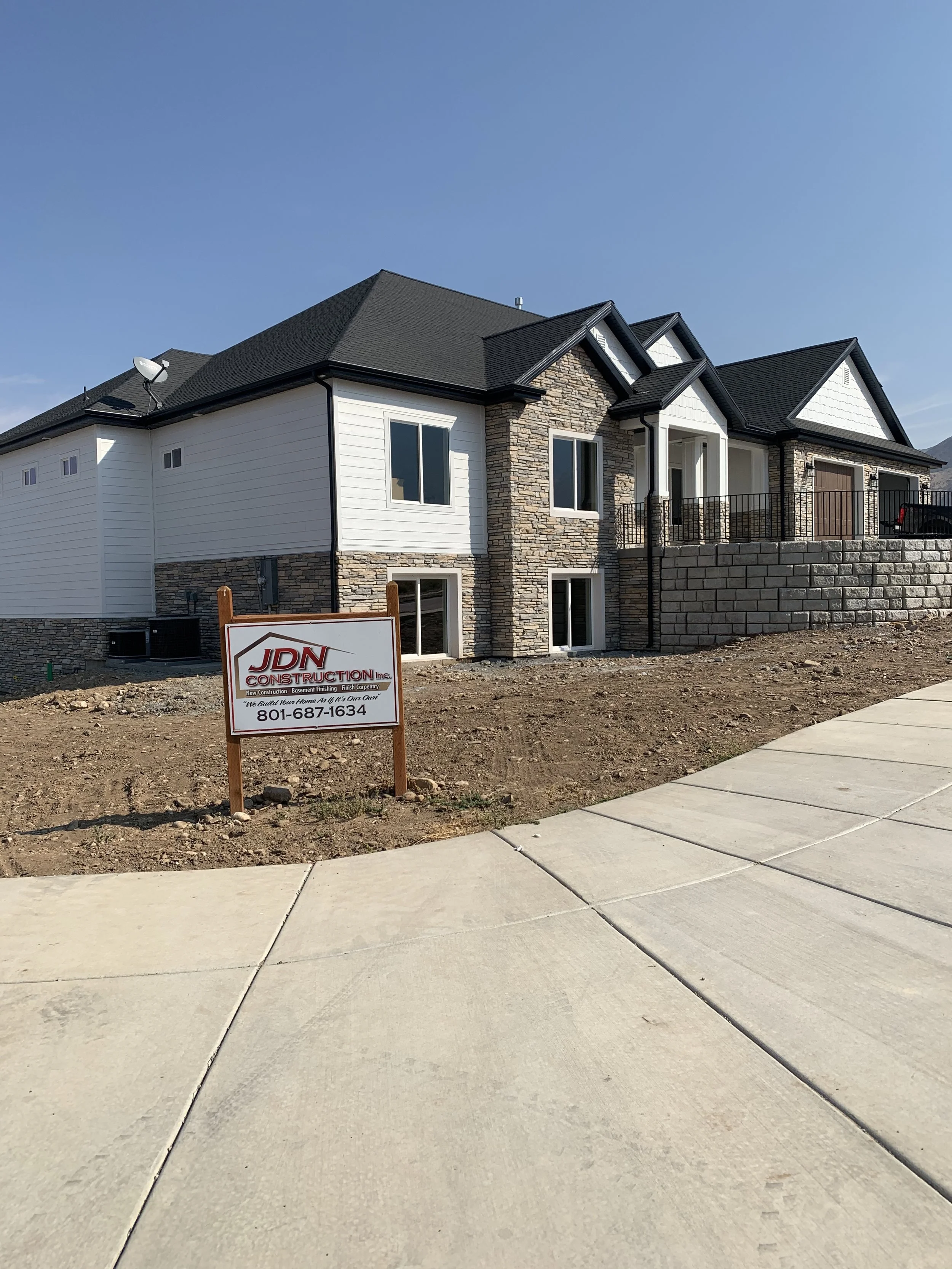 Newly constructed house with white siding, stone accents, and a black roof, situated behind a concrete sidewalk, with a sign from JDN Construction in the front.