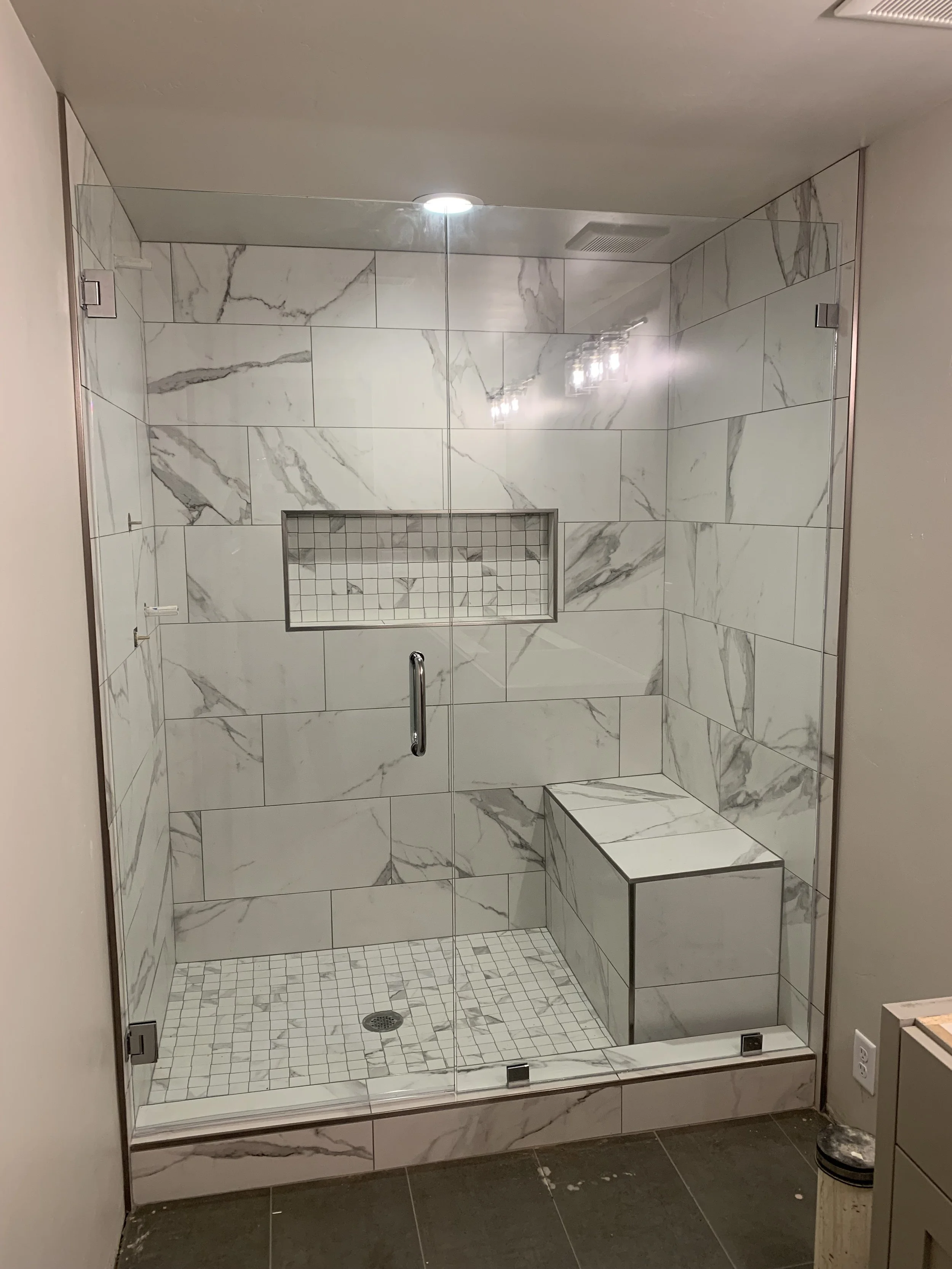 A modern walk-in shower with glass doors, large marble tiles, a built-in marble bench, a tiled niche for toiletries, and a floor drain, with a partial view of the surrounding bathroom.