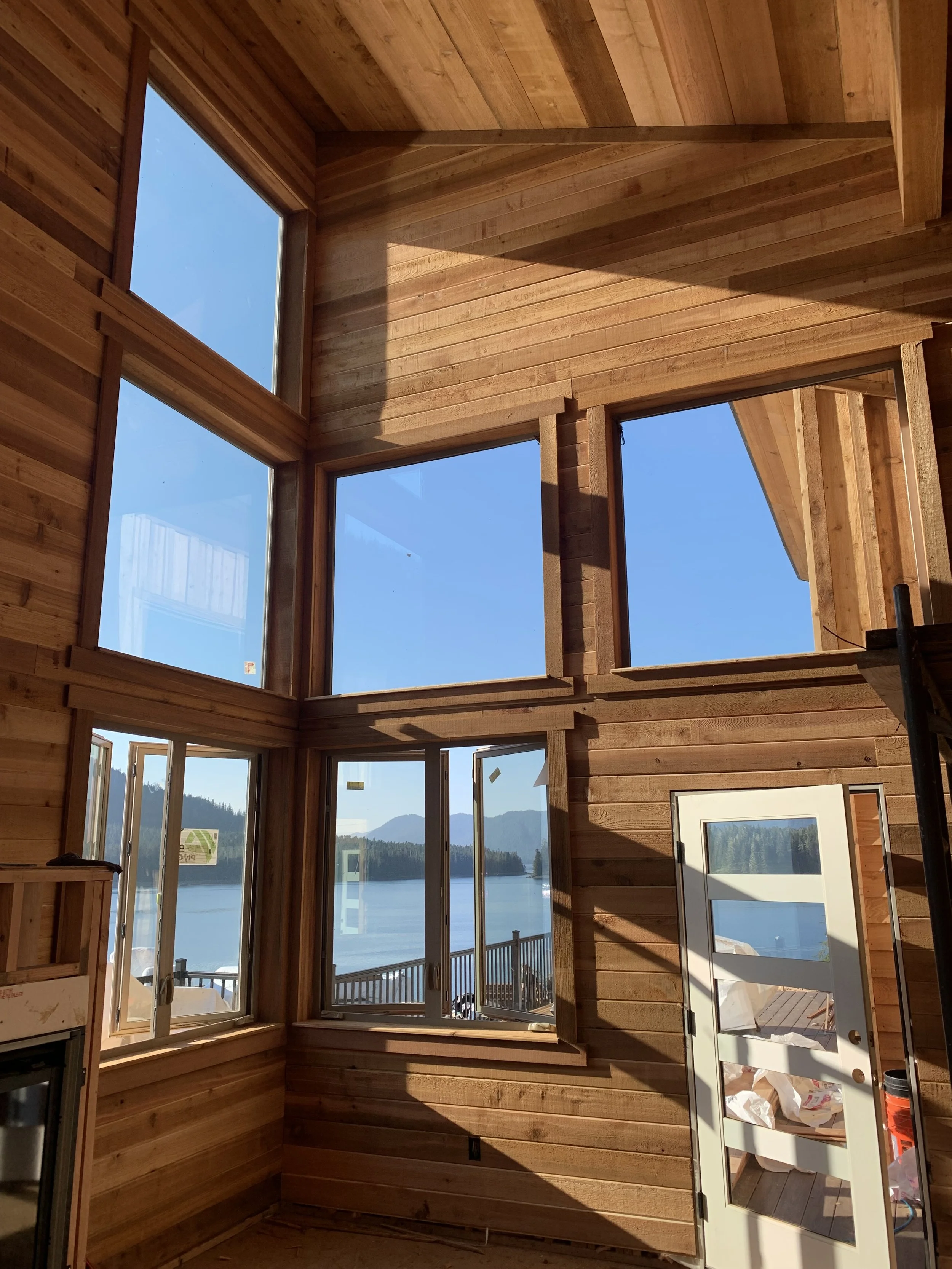 Interior of a wooden house under construction with large windows and a view of water and mountains outside.