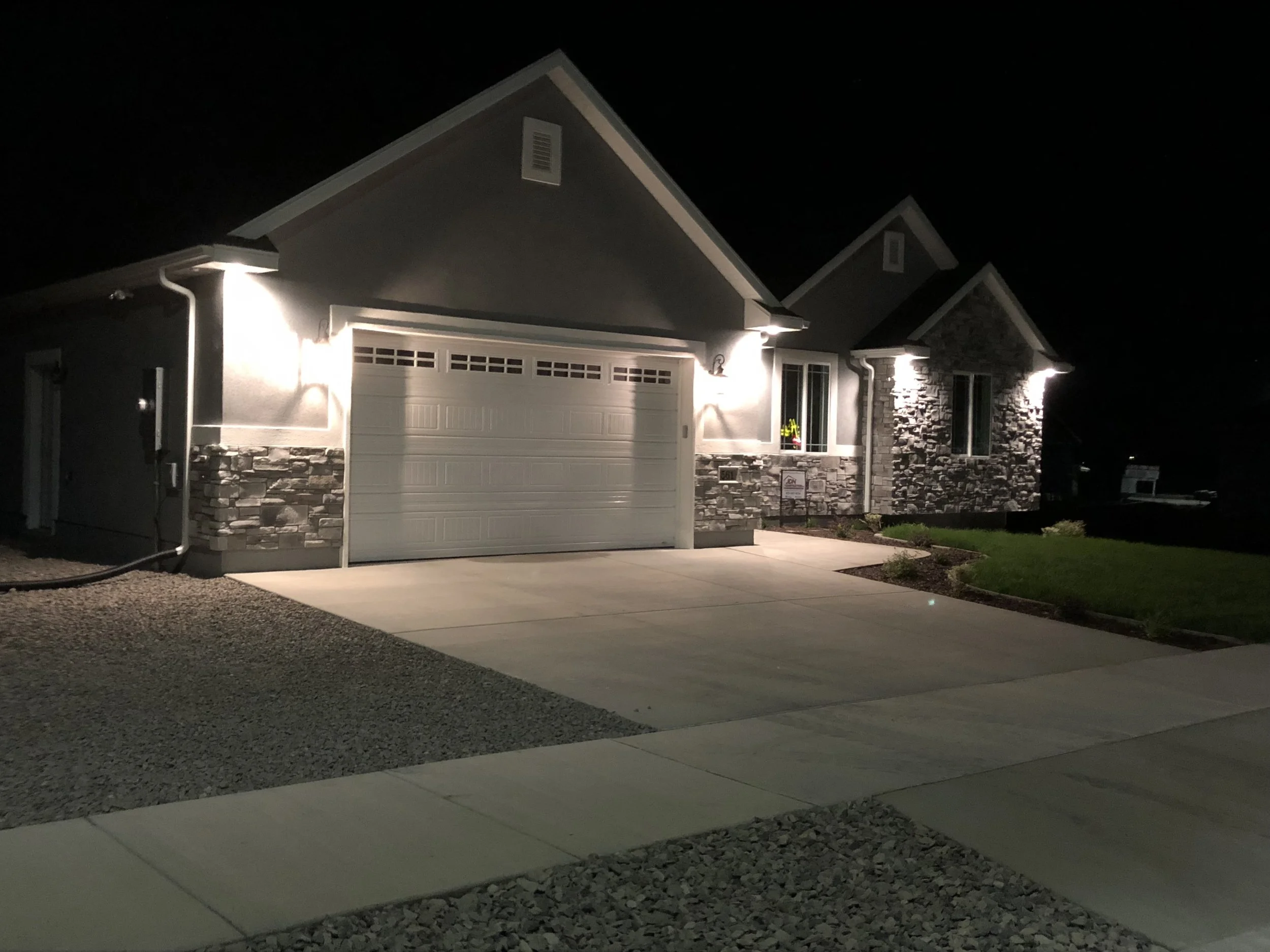 Night view of a house with a stone and siding exterior, illuminated by exterior lights. The house has a two-car garage, a concrete driveway, and a small front lawn with landscaping.
