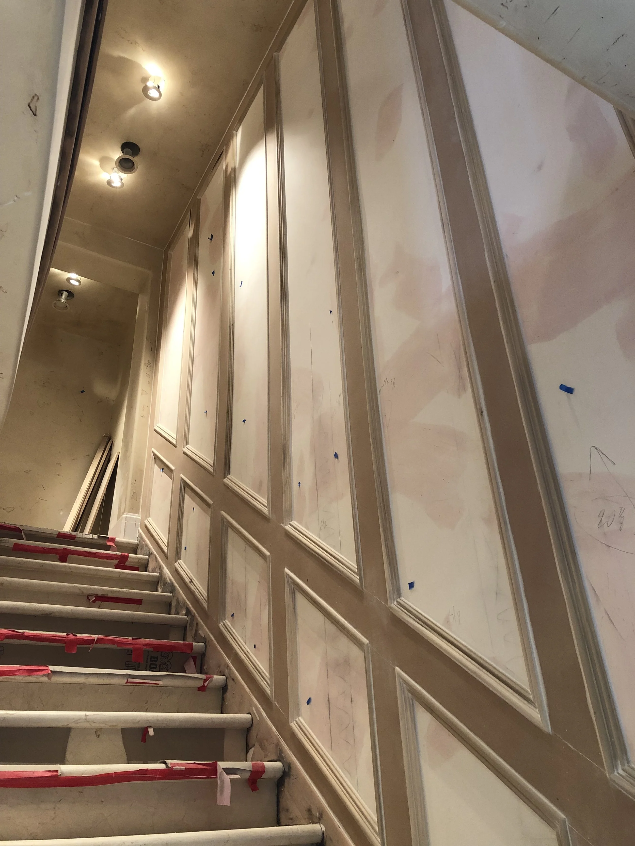 Interior view of a staircase under construction, with unfinished wall paneling and lighting fixtures.