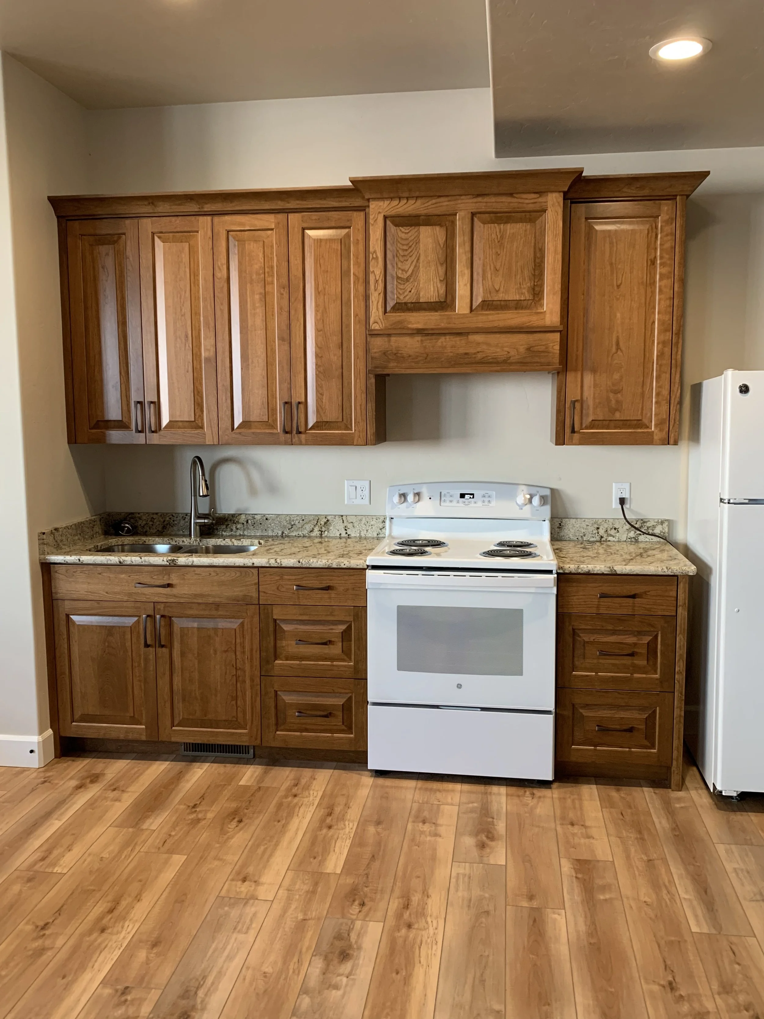 A kitchen with wooden cabinets, granite countertops, a white electric stove, a sink with a faucet, and a refrigerator on the right.