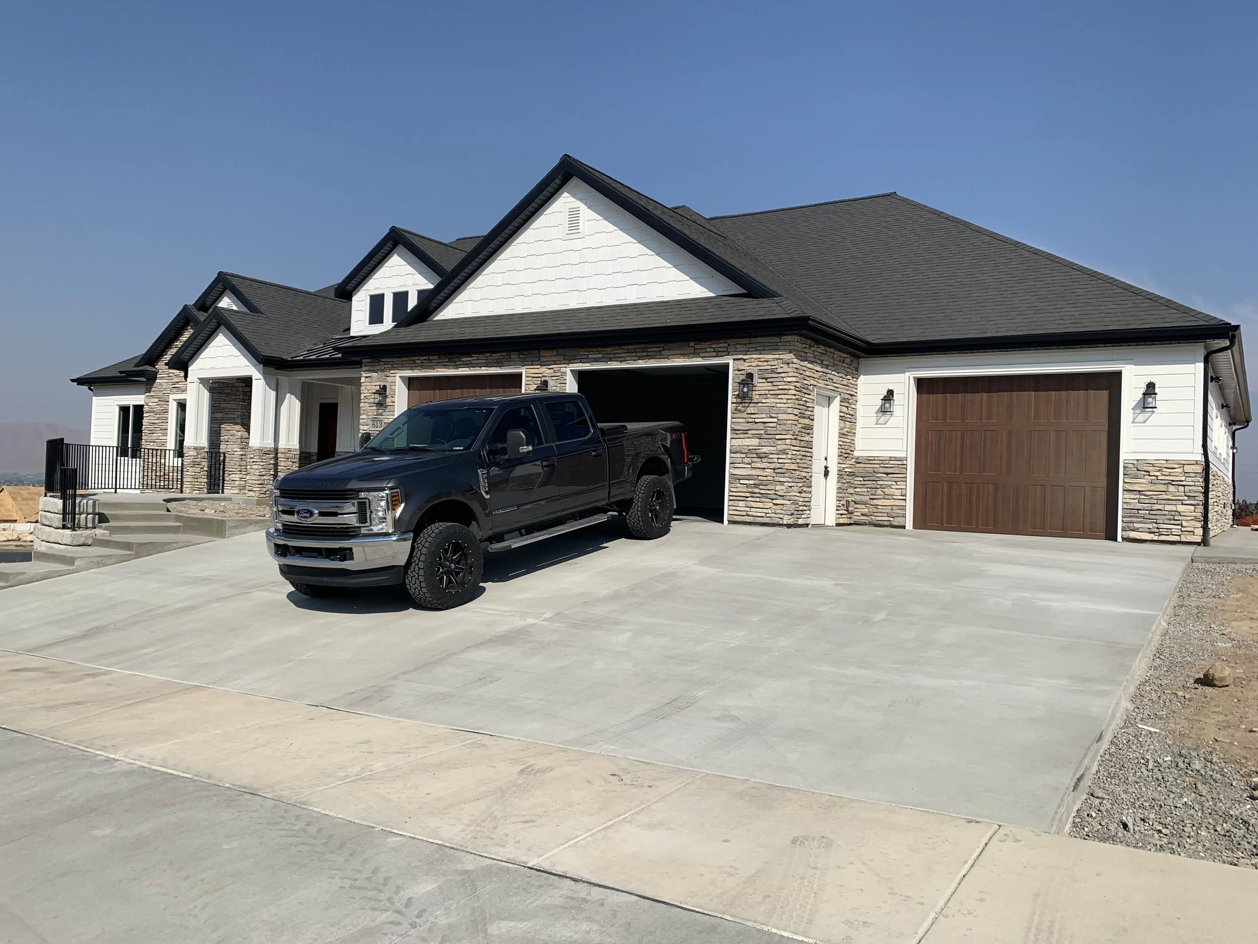 A modern two-story house with a black pickup truck parked in the driveway, featuring a stone and white siding exterior, a dark shingle roof, and a wooden garage door. The house has steps leading up to the front door and black exterior light fixtures.
