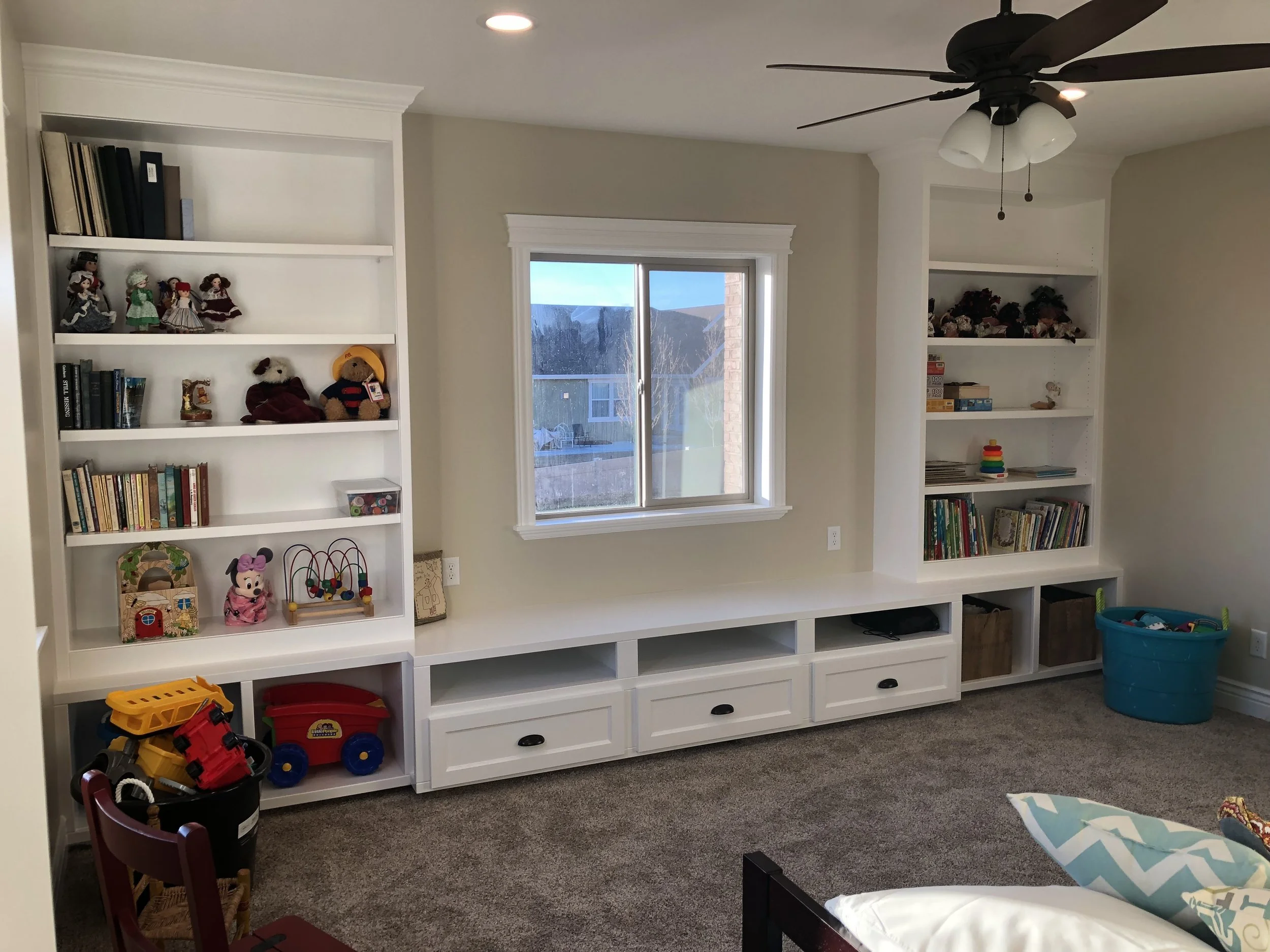 Living room with two white built-in bookshelves filled with toys, books, and decorations, a window in the center, a ceiling fan with light fixtures, a blue storage bin filled with toys, and a carpeted floor.