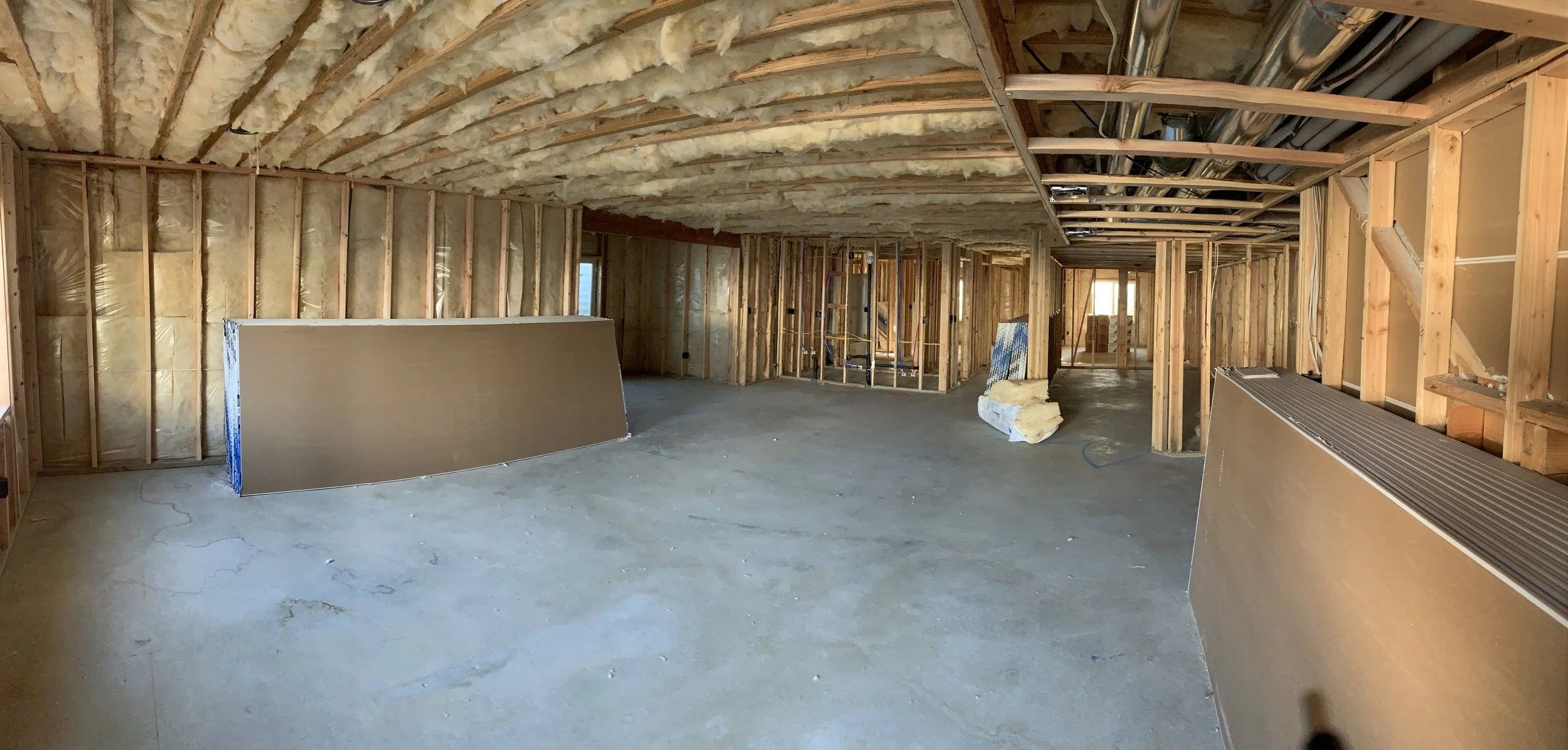 Interior view of a house under construction, showing exposed wall studs, insulation, and ceiling framework with ductwork, with construction materials on the floor.